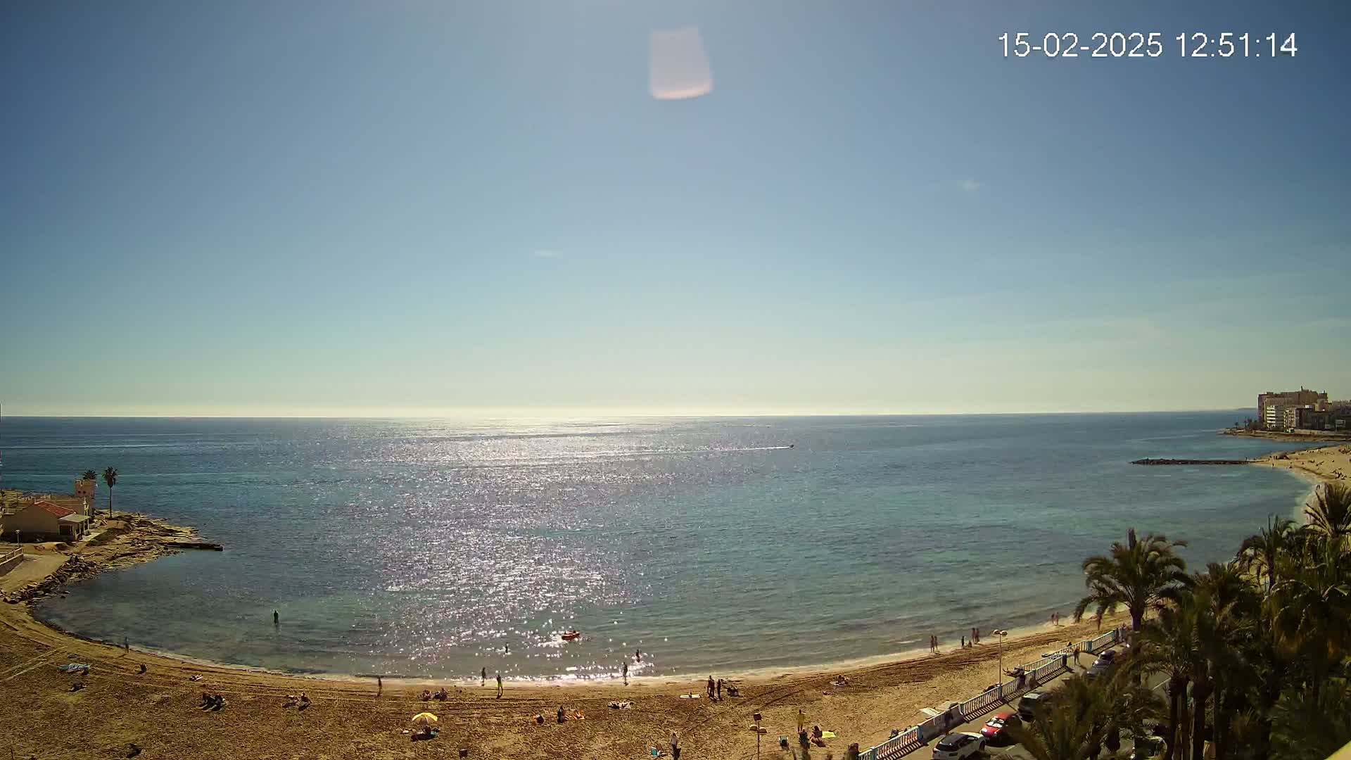 A sunny day at a sandy beach with calm, sparkling water, showing a few people on the beach and in the water, and palm trees along the shore.