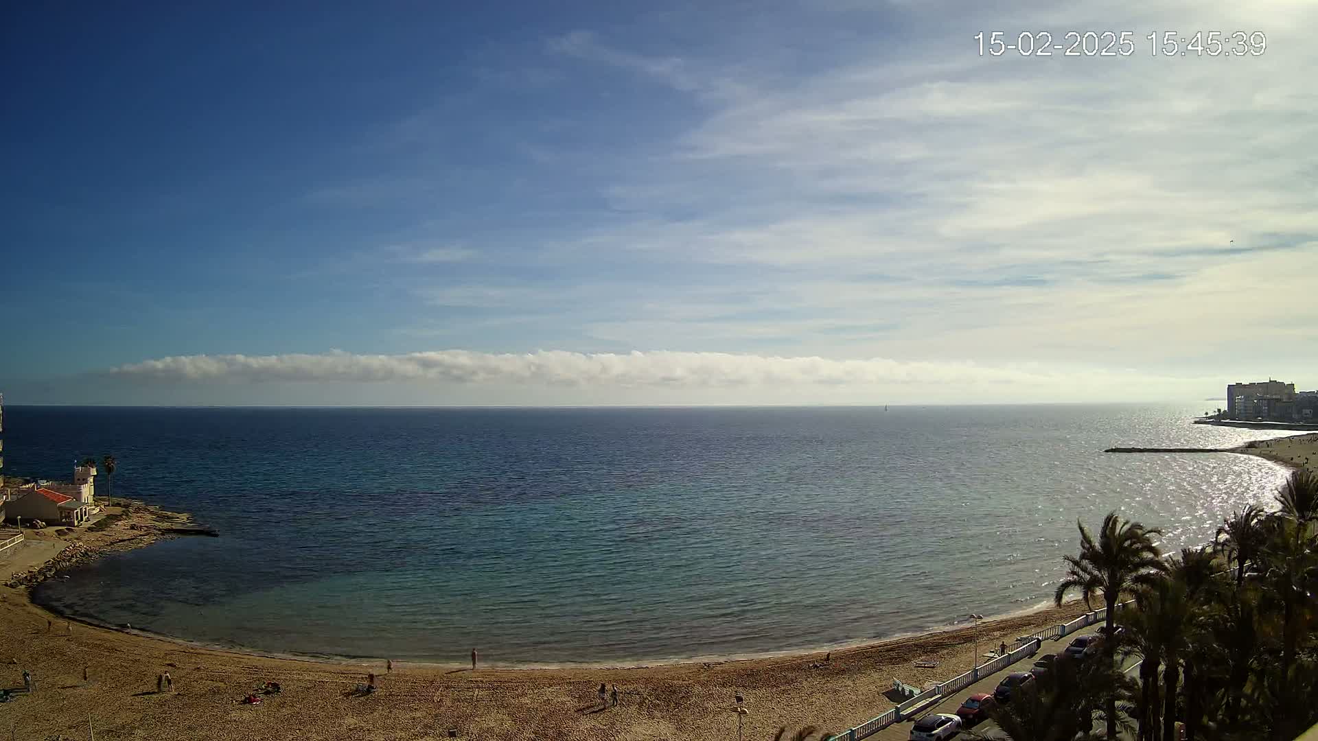 A sandy beach curves around a calm, blue-green sea under a mostly sunny sky with a thin band of clouds on the horizon.