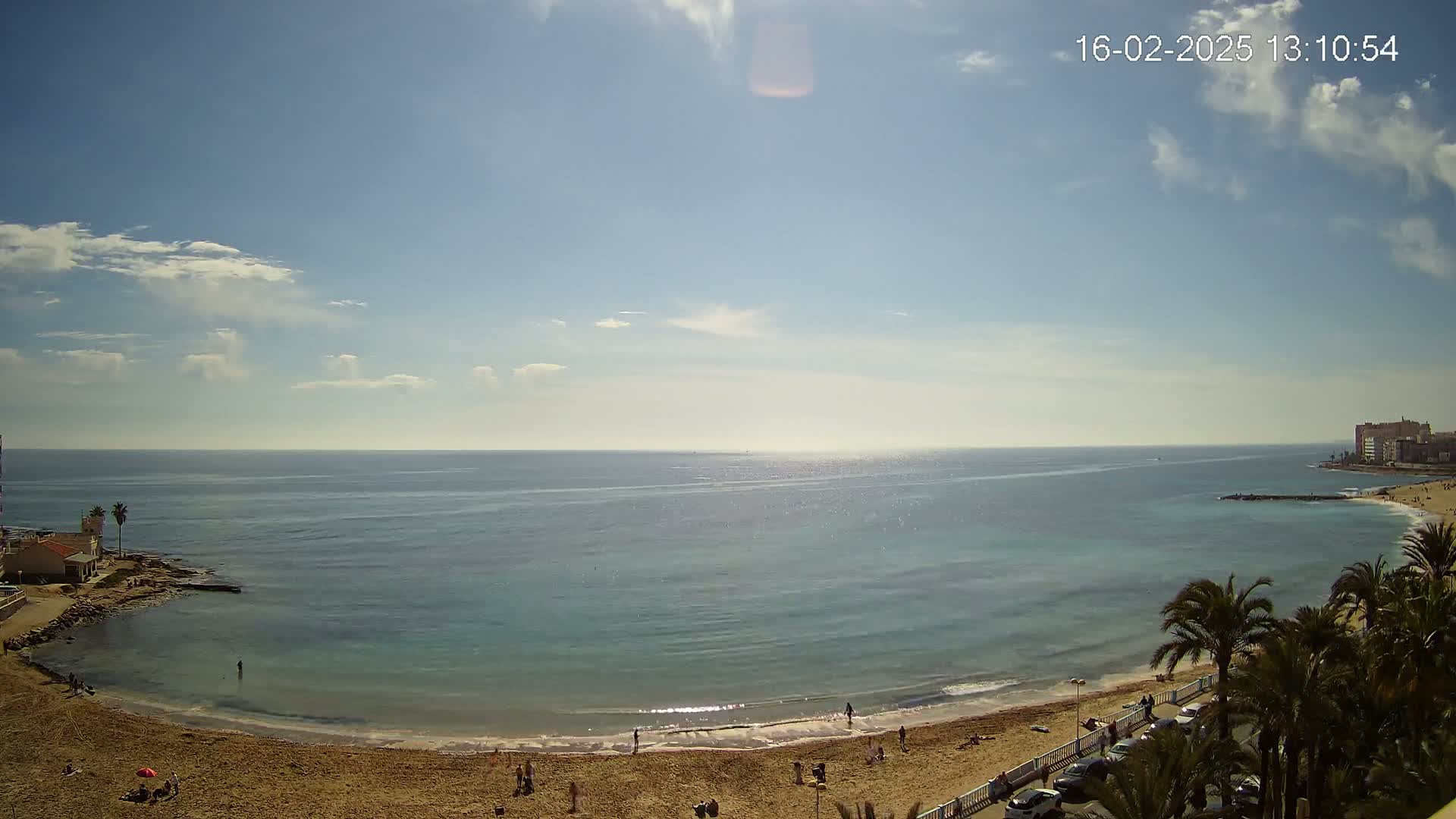 A sandy beach curves around a calm, turquoise sea under a mostly sunny sky with a few scattered clouds.