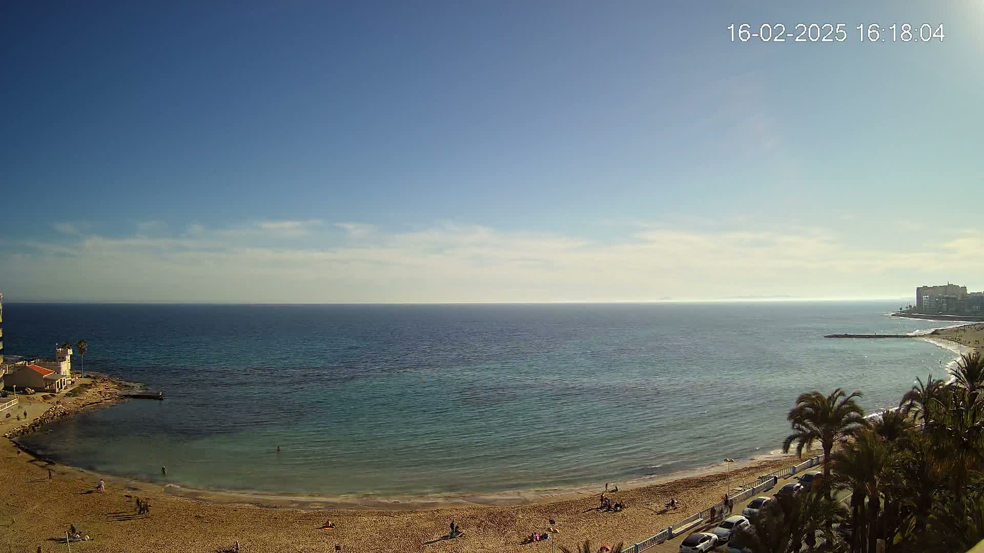 A sandy beach curves around a calm, teal-colored ocean under a mostly clear blue sky with a few scattered clouds.