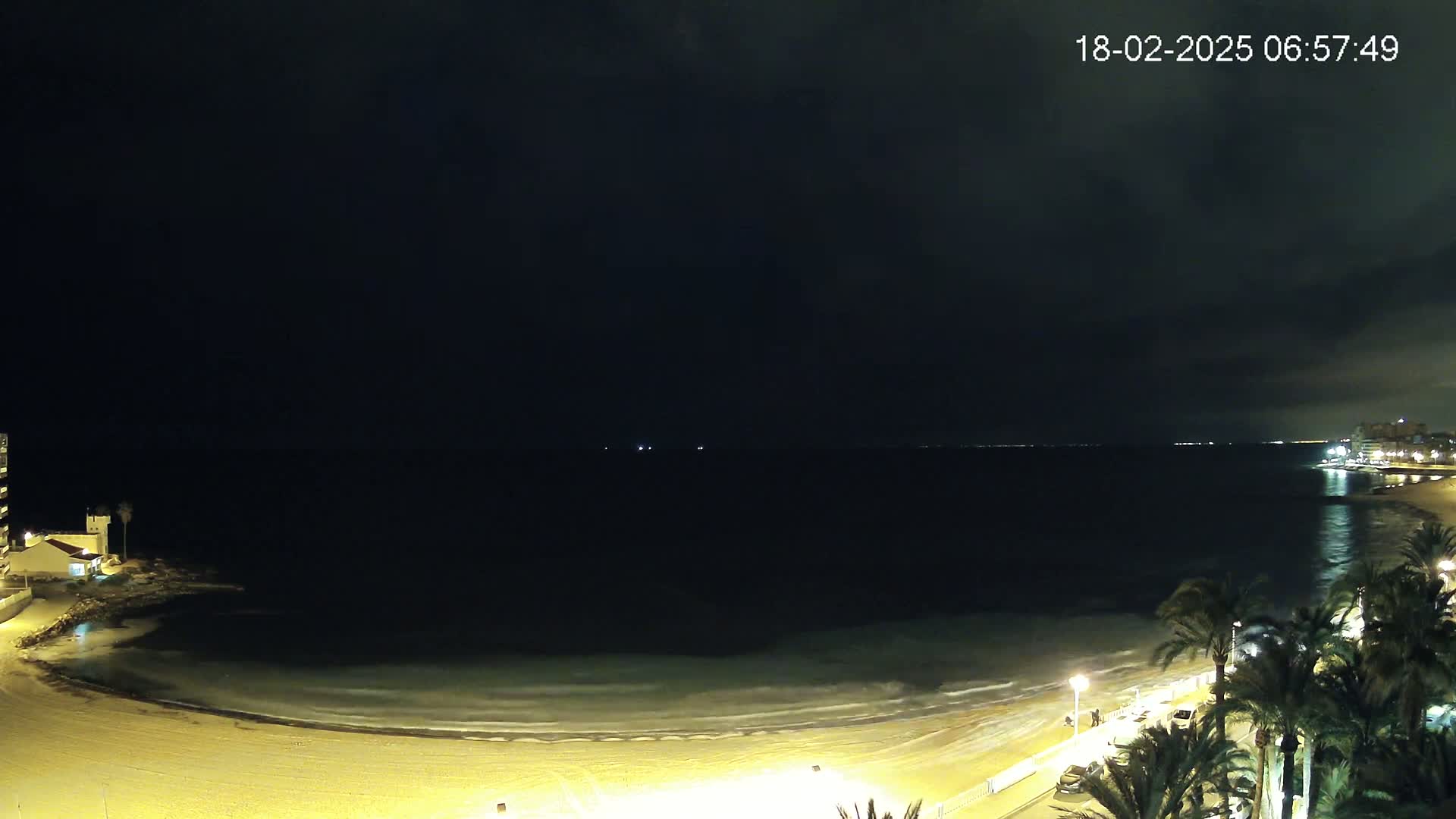 A dark, nighttime view of a beach with gentle waves lapping the shore, illuminated by lights along a palm tree-lined promenade.