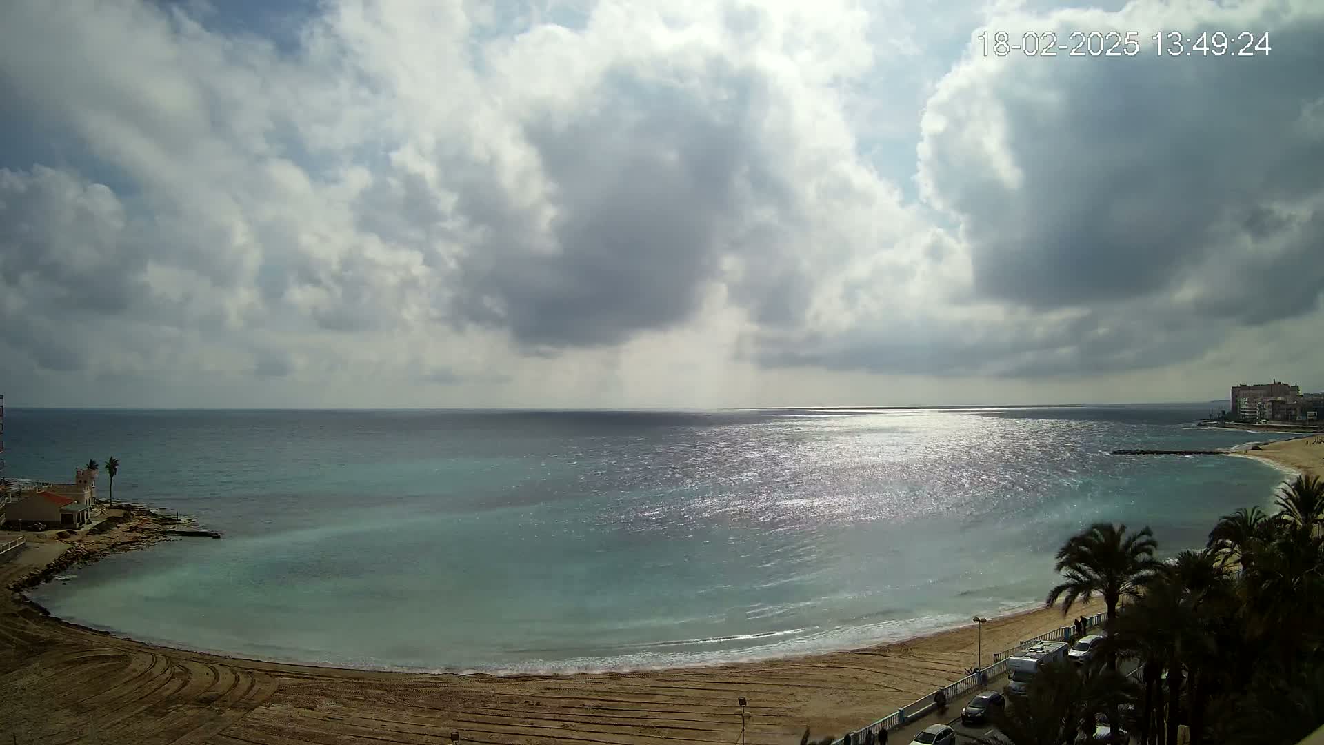 A partly cloudy sky overlooks a sandy beach and calm, sparkling turquoise sea, with palm trees and buildings visible in the distance.