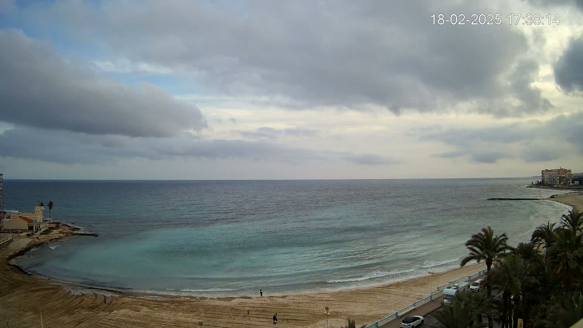 A sandy beach curves along a calm, teal sea under a mostly cloudy sky.