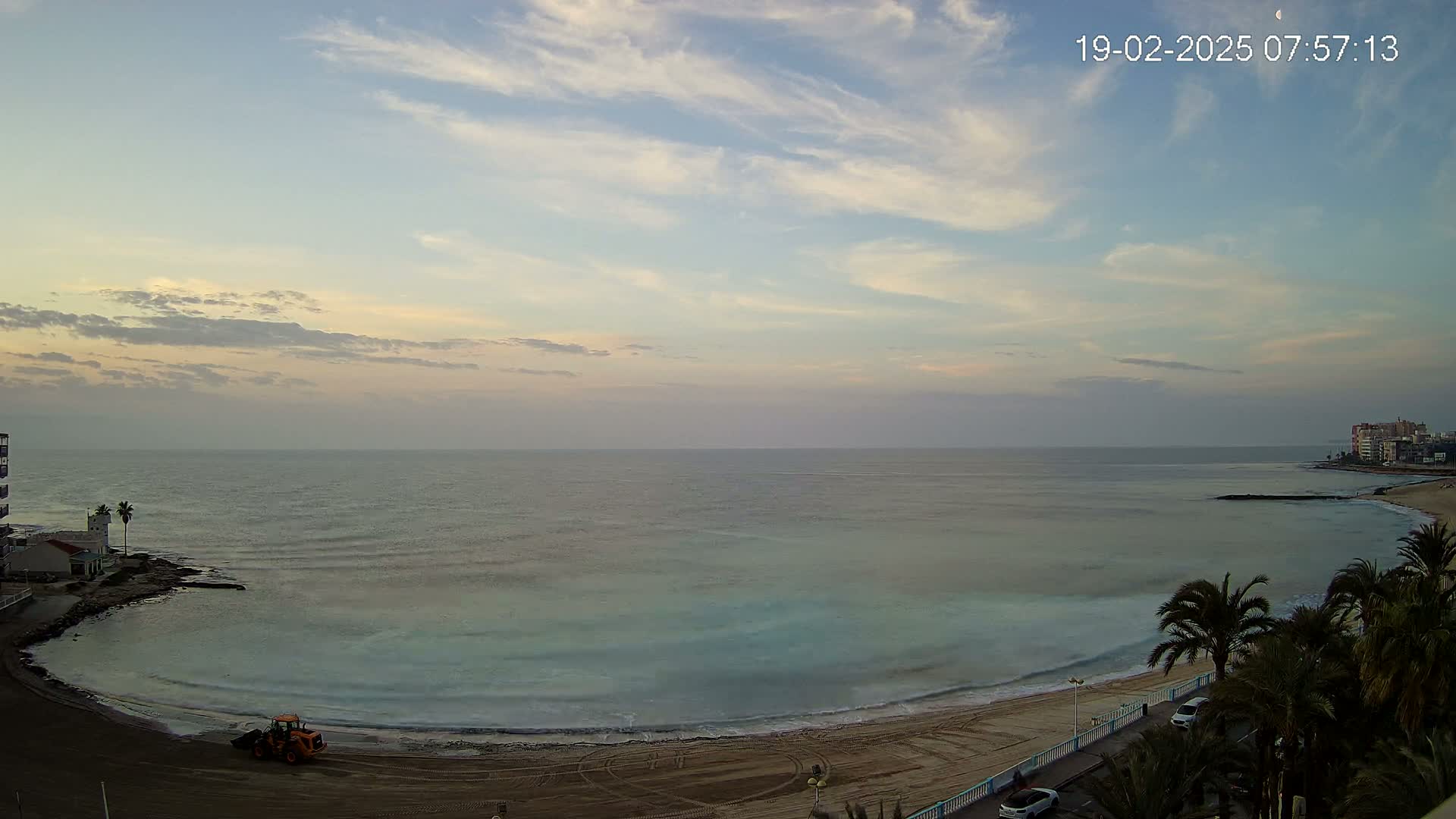 A tranquil beach scene at dawn under a mostly clear sky shows a calm sea with gentle waves lapping a sandy shore where a small bulldozer is parked.