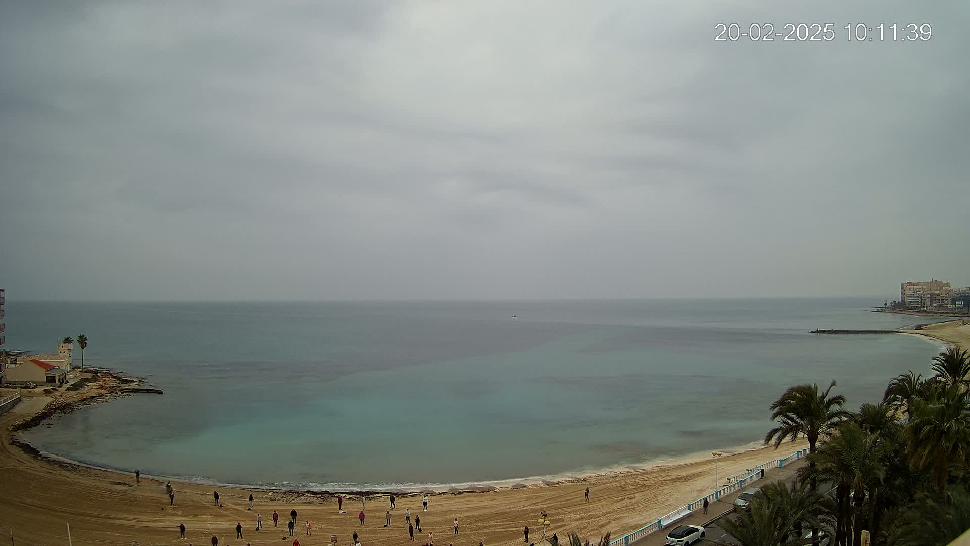 A mostly cloudy day shows a sandy beach curving around a calm, teal-colored bay with several people on the sand and palm trees lining the shore.