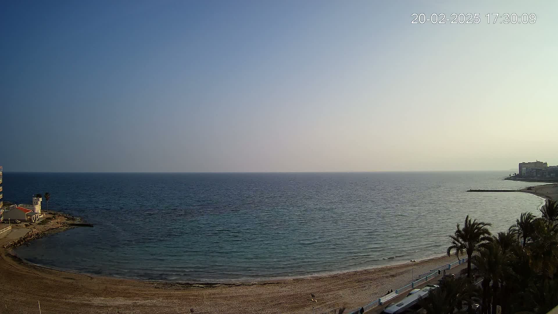 A calm, clear day reveals a sandy beach curving into a calm sea, with buildings visible in the distance and palm trees in the foreground.