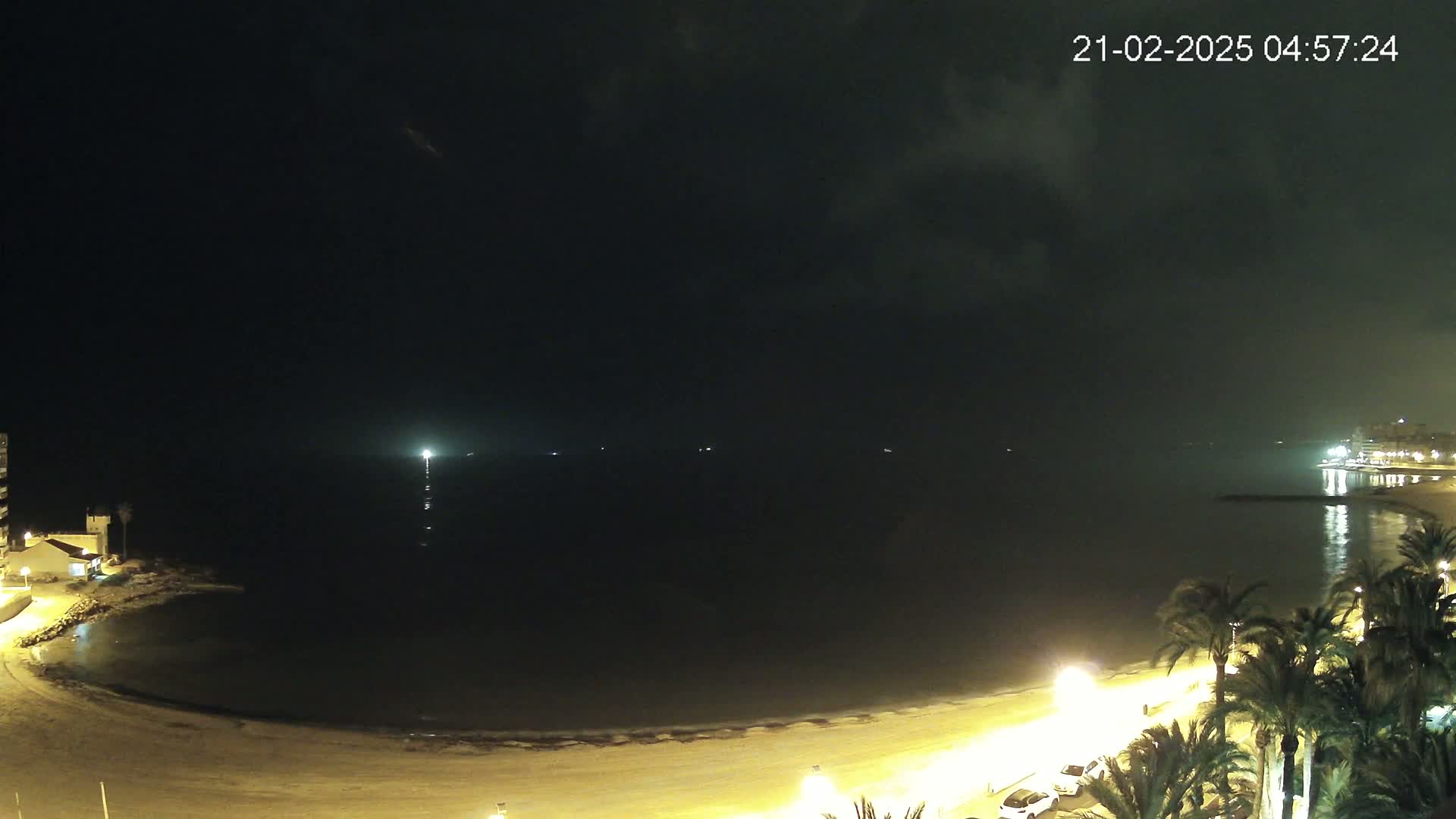 A nighttime view shows a dark, calm sea lapping a sandy beach lined with palm trees and buildings under a mostly cloudy sky.
