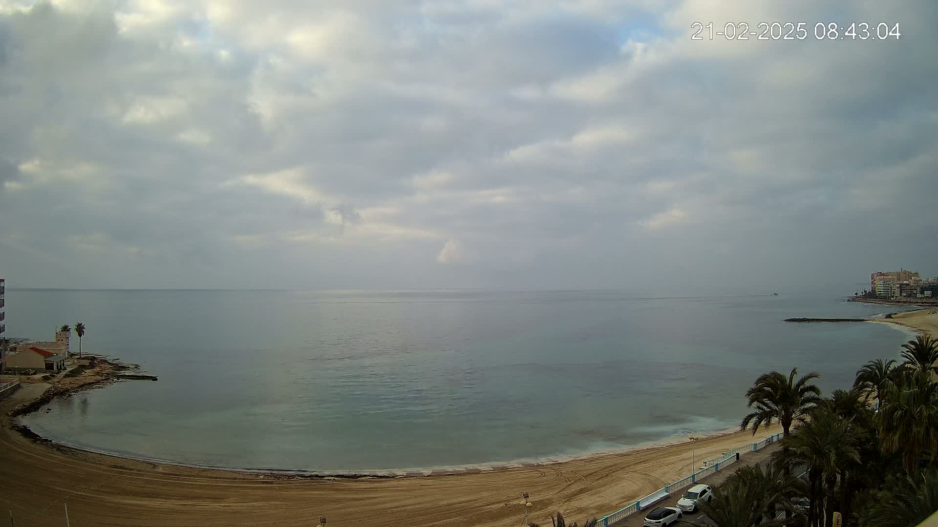 A mostly cloudy day overlooks a calm, teal ocean meeting a sandy beach with palm trees and buildings in the distance.