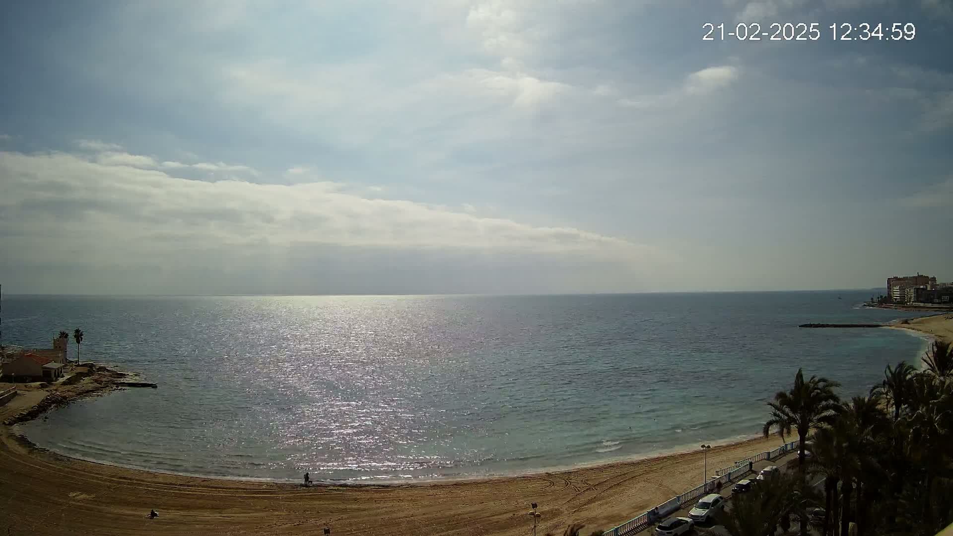 A sandy beach curves around a calm, sunlit sea under a partly cloudy sky.