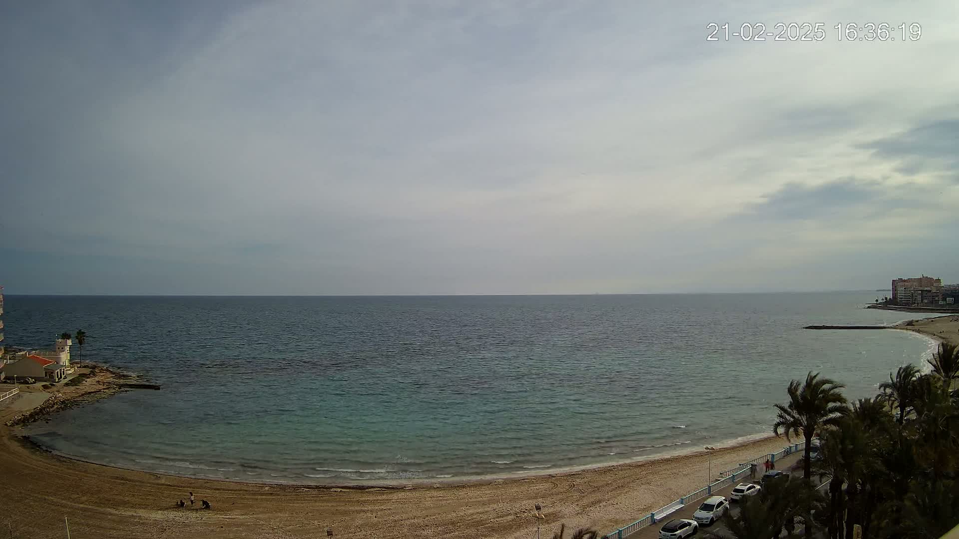 A sandy beach curves around a calm, teal sea under a mostly cloudy sky.