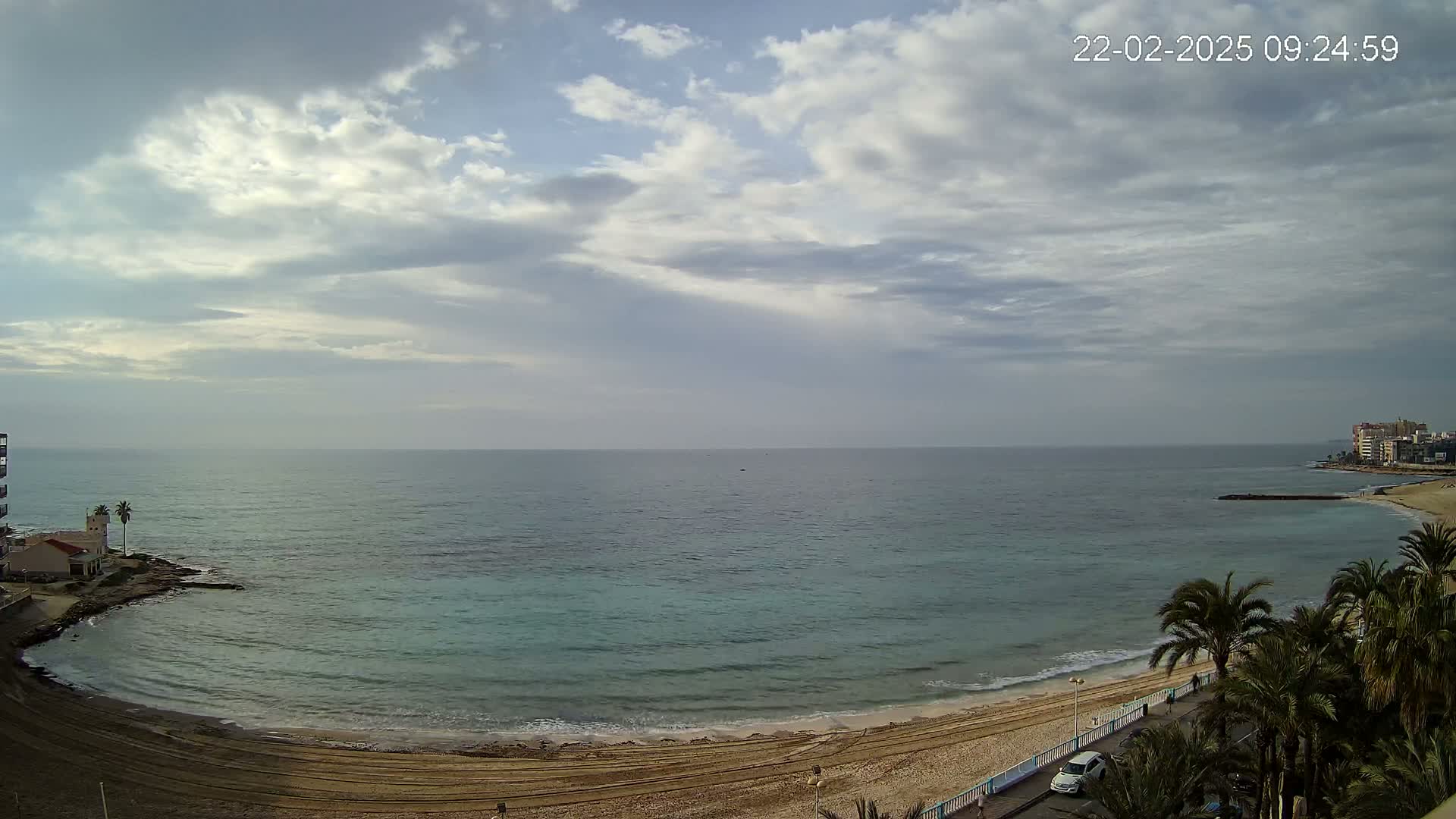 A partly cloudy sky overlooks a calm, teal ocean meeting a sandy beach lined with palm trees and buildings.