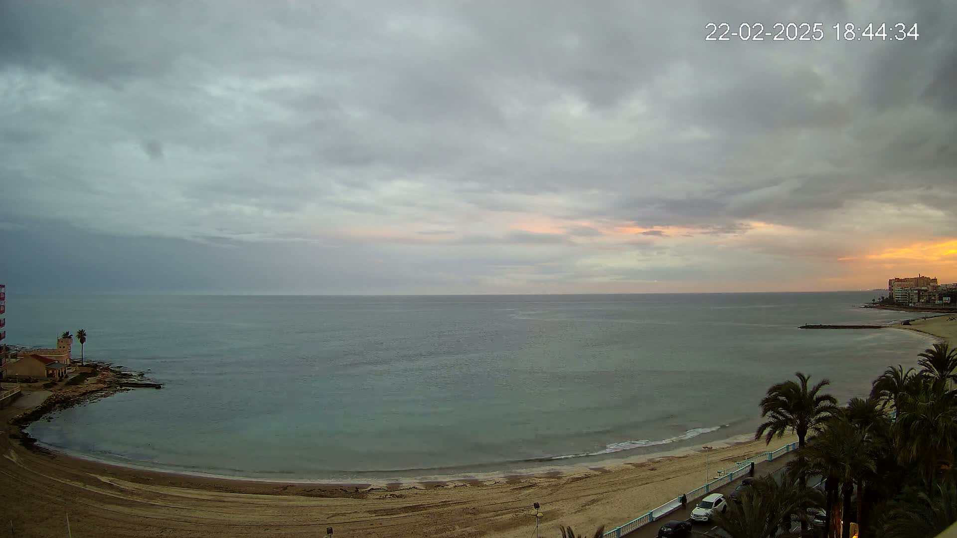 A sandy beach curves along a calm sea under a cloudy sky with hints of sunset.