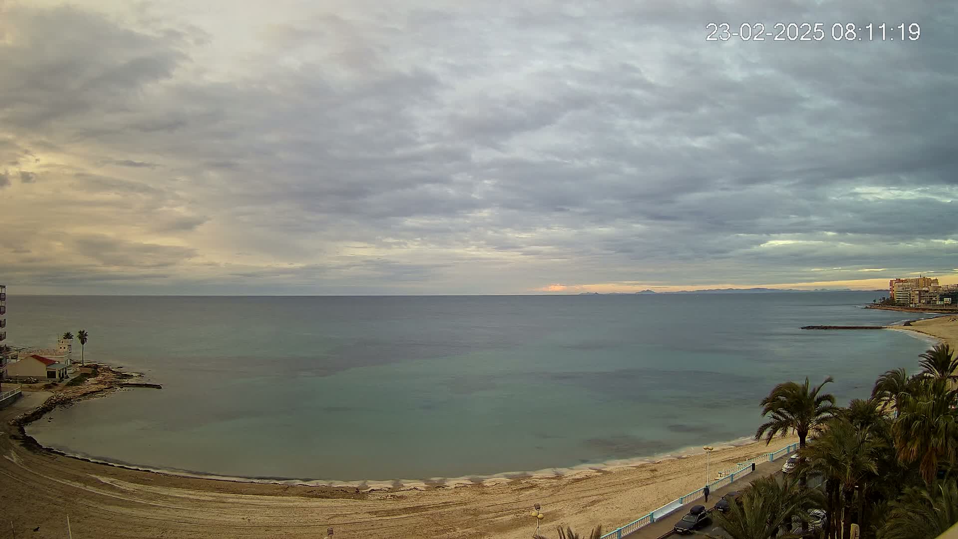A sandy beach curves along a calm, teal sea under a mostly cloudy, overcast sky.