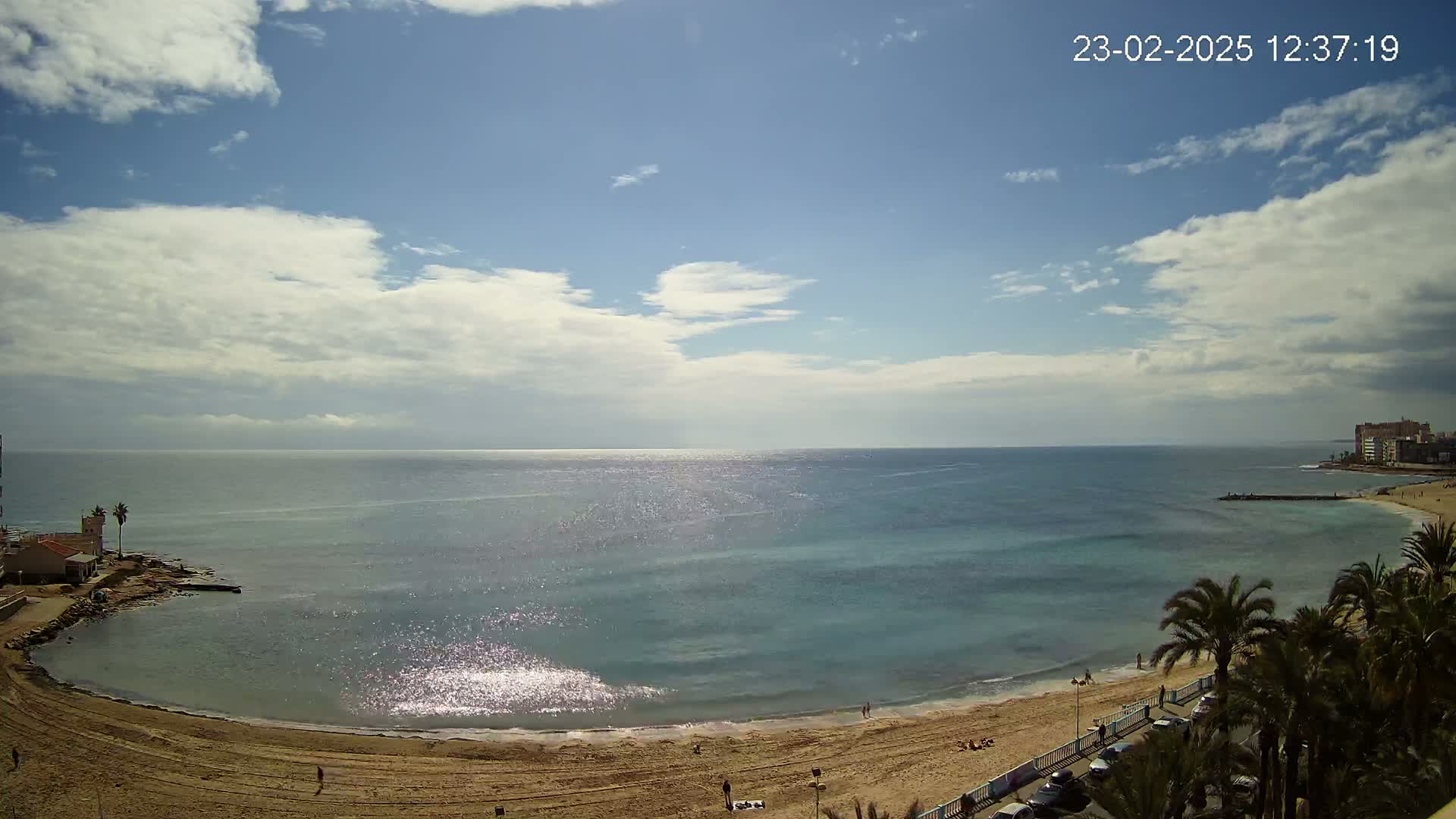 A partly cloudy day shows a sandy beach curving around a calm, sparkling blue sea, with palm trees and buildings visible in the distance.