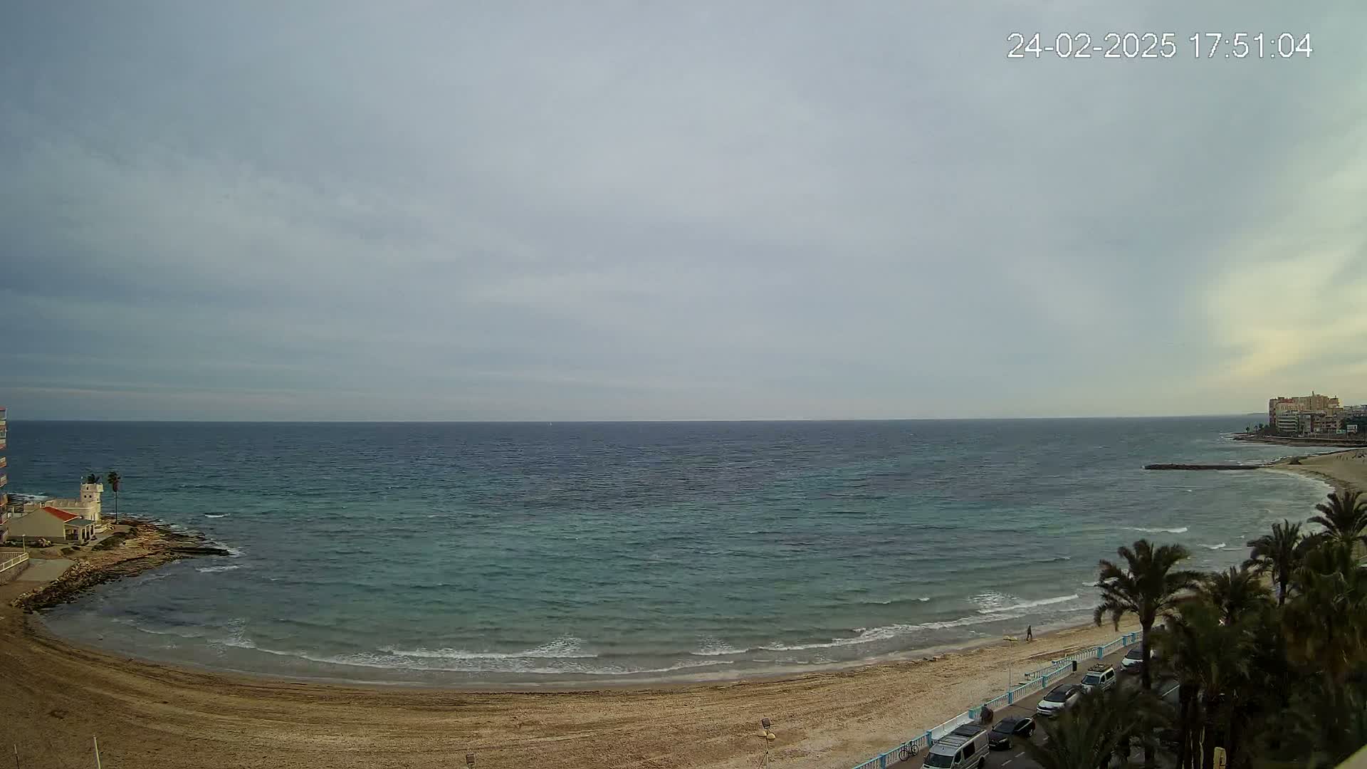 A sandy beach curves around a calm, teal-colored sea under an overcast sky.