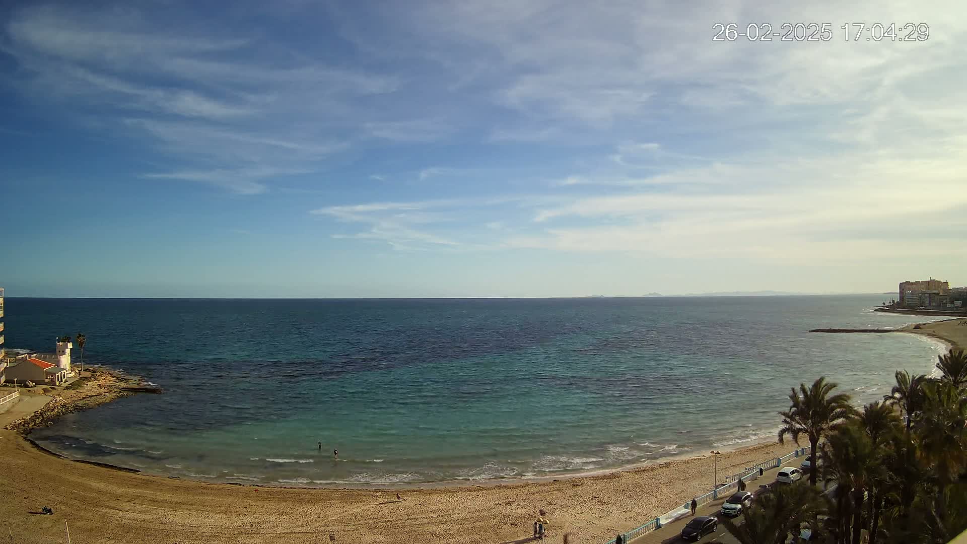 A sandy beach curves around a calm, teal-colored sea under a mostly clear sky with scattered clouds.