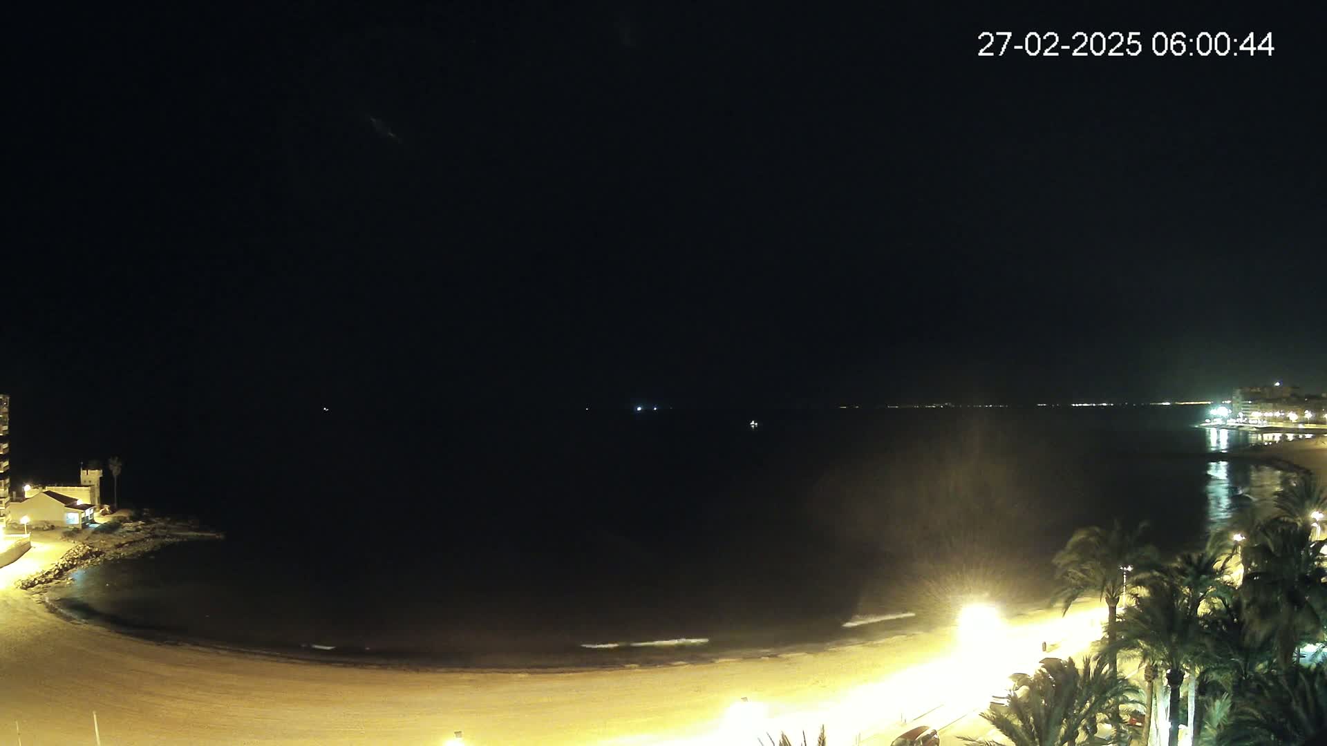 A dark night scene shows a beach illuminated by artificial lights, with calm water and palm trees lining the shore.