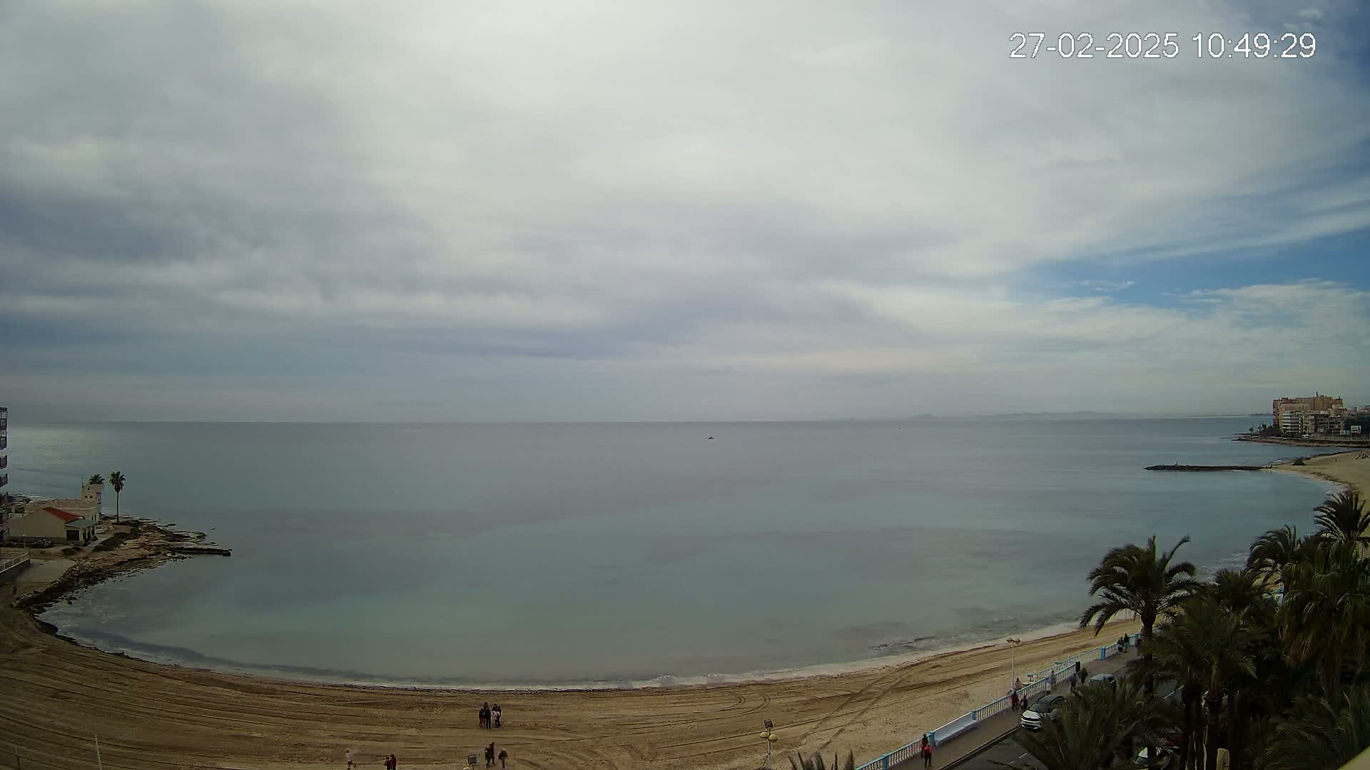 A sandy beach curves around a calm, light-blue sea under a mostly cloudy sky.