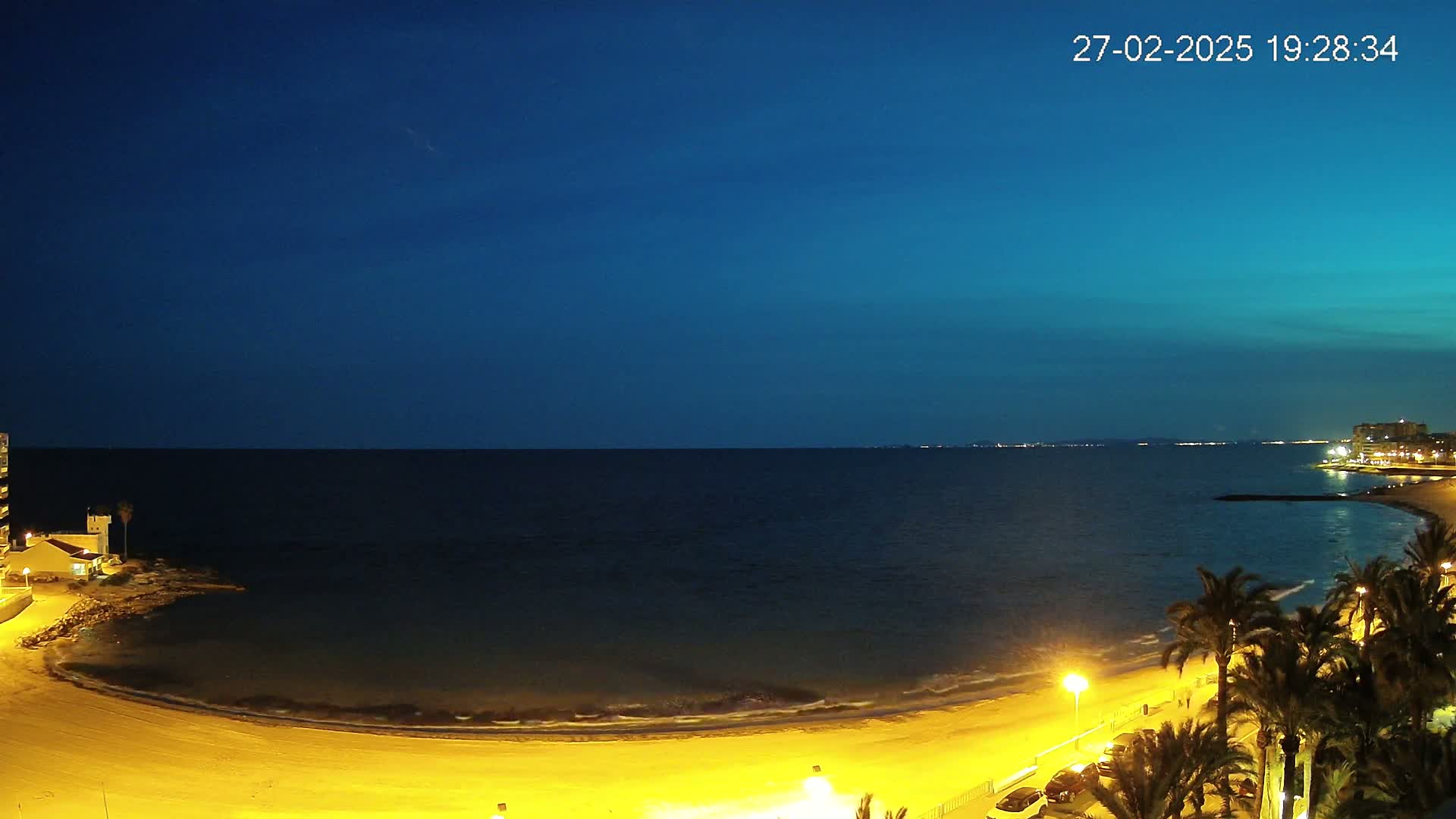 A crescent-shaped beach at night is illuminated by artificial lights, bordered by palm trees and buildings, overlooking a dark blue sea under a twilight sky.
