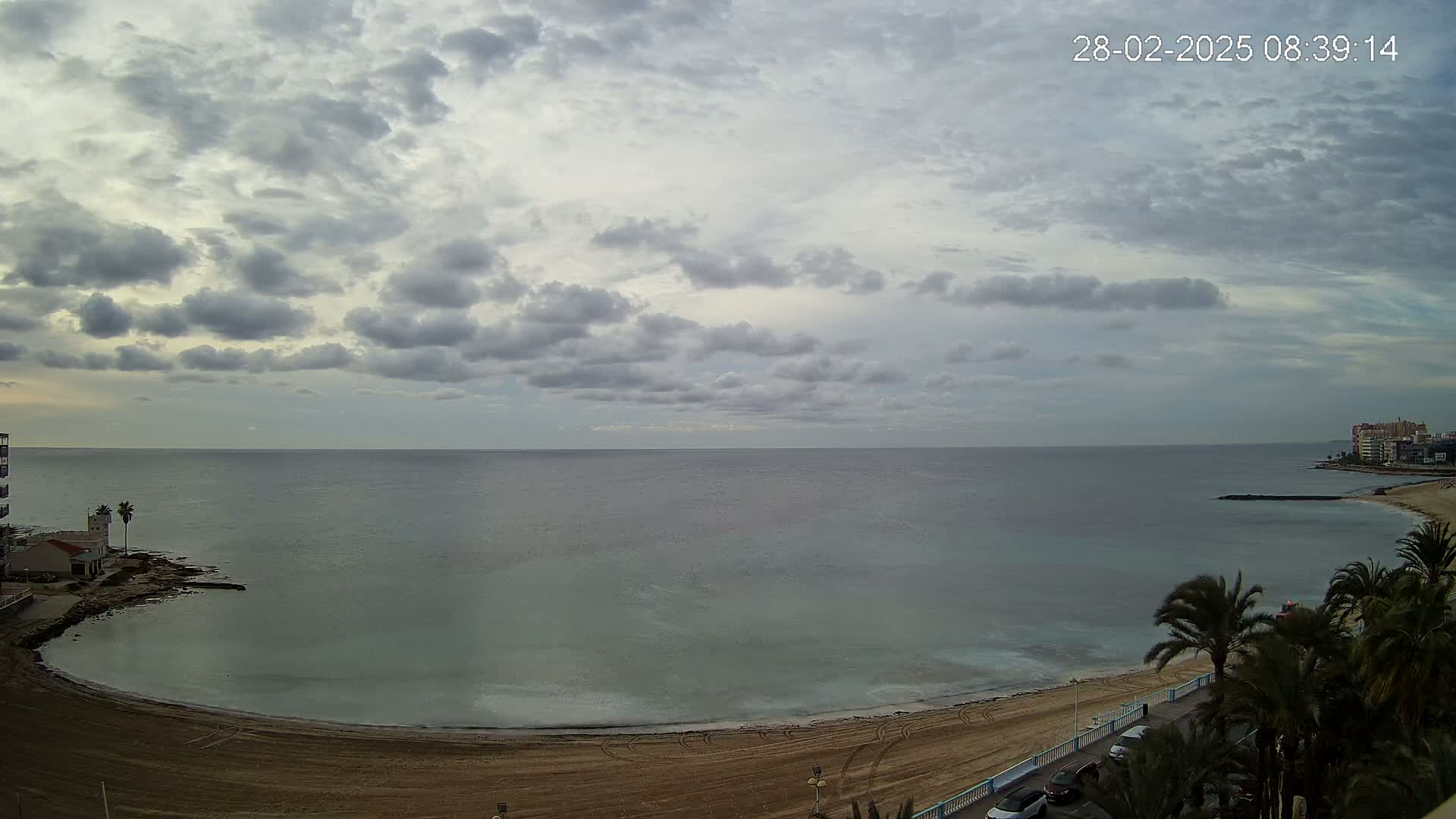 A mostly cloudy sky overlooks a calm sea curving into a sandy beach lined with palm trees and buildings.