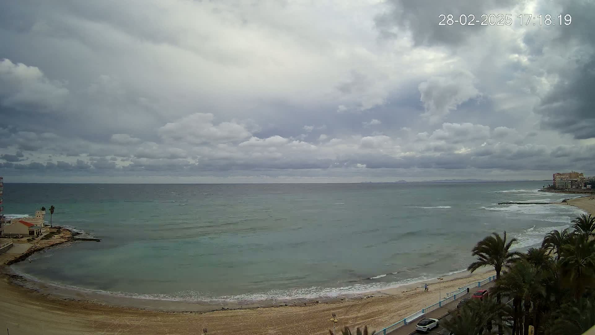 A mostly cloudy day shows a view of a sandy beach, calm ocean waves, and palm trees lining a road next to the beach.
