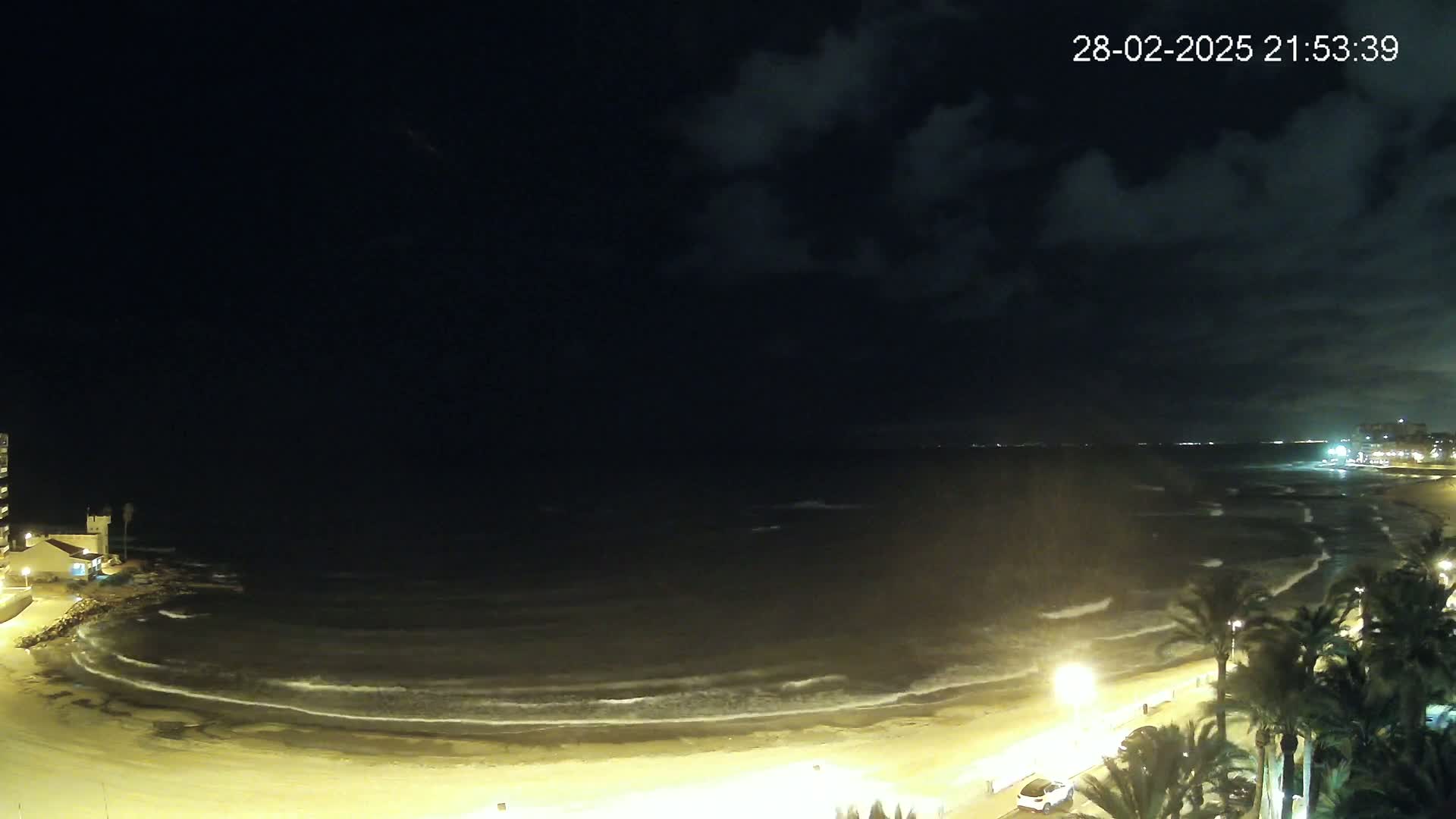 A nighttime beach scene shows gently rolling waves lapping on a sandy shore, illuminated by streetlights, under a partly cloudy sky.
