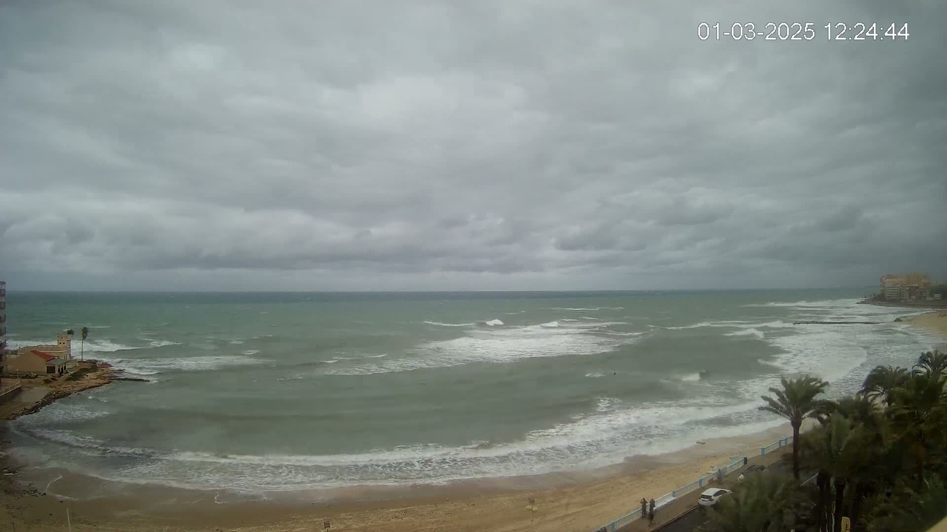 A mostly cloudy day shows a beach with moderate waves and palm trees lining a road next to the sand.
