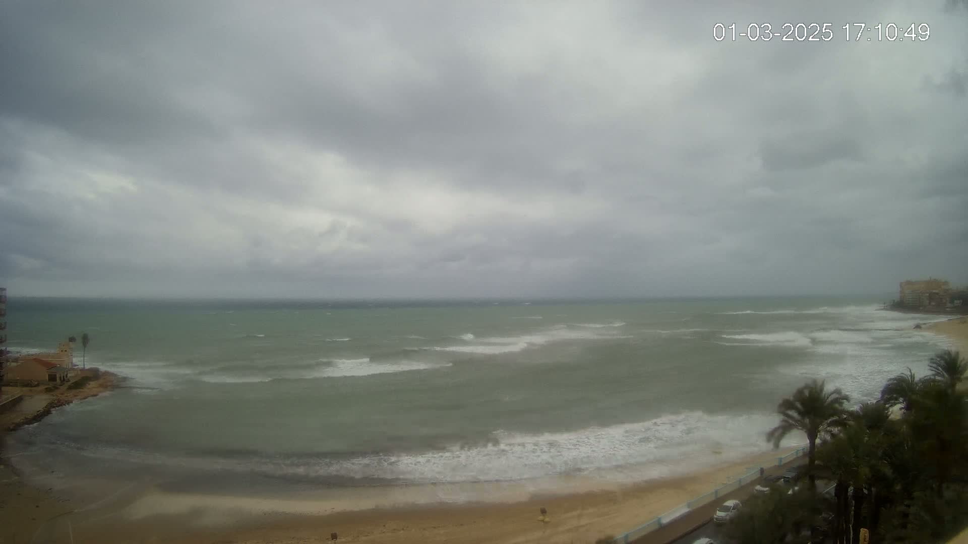A windy, overcast day at a beach shows choppy waves breaking on the shore near buildings and palm trees.