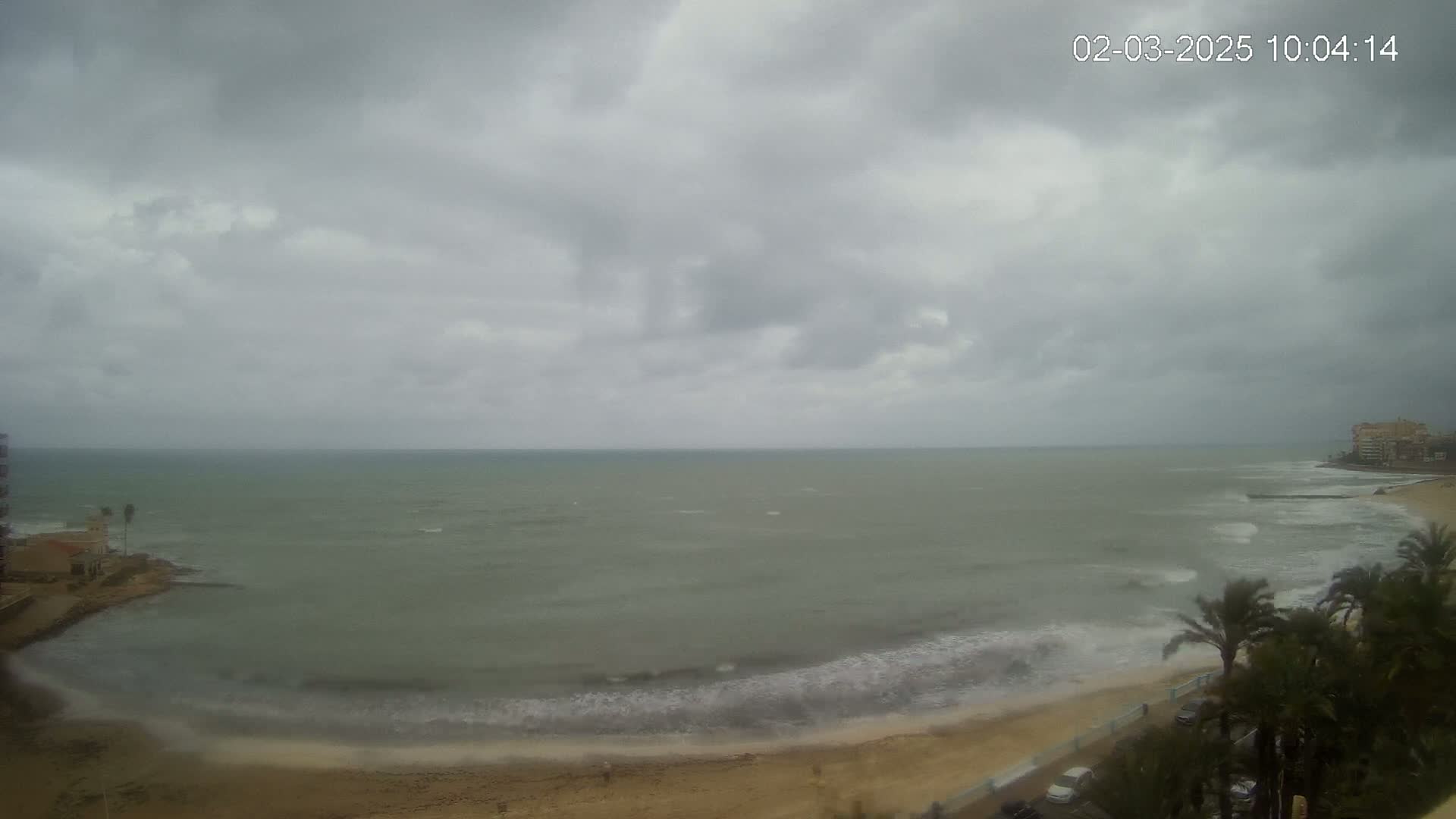 A mostly cloudy day shows a gray ocean with small waves breaking on a sandy beach, palm trees, and buildings in the distance.