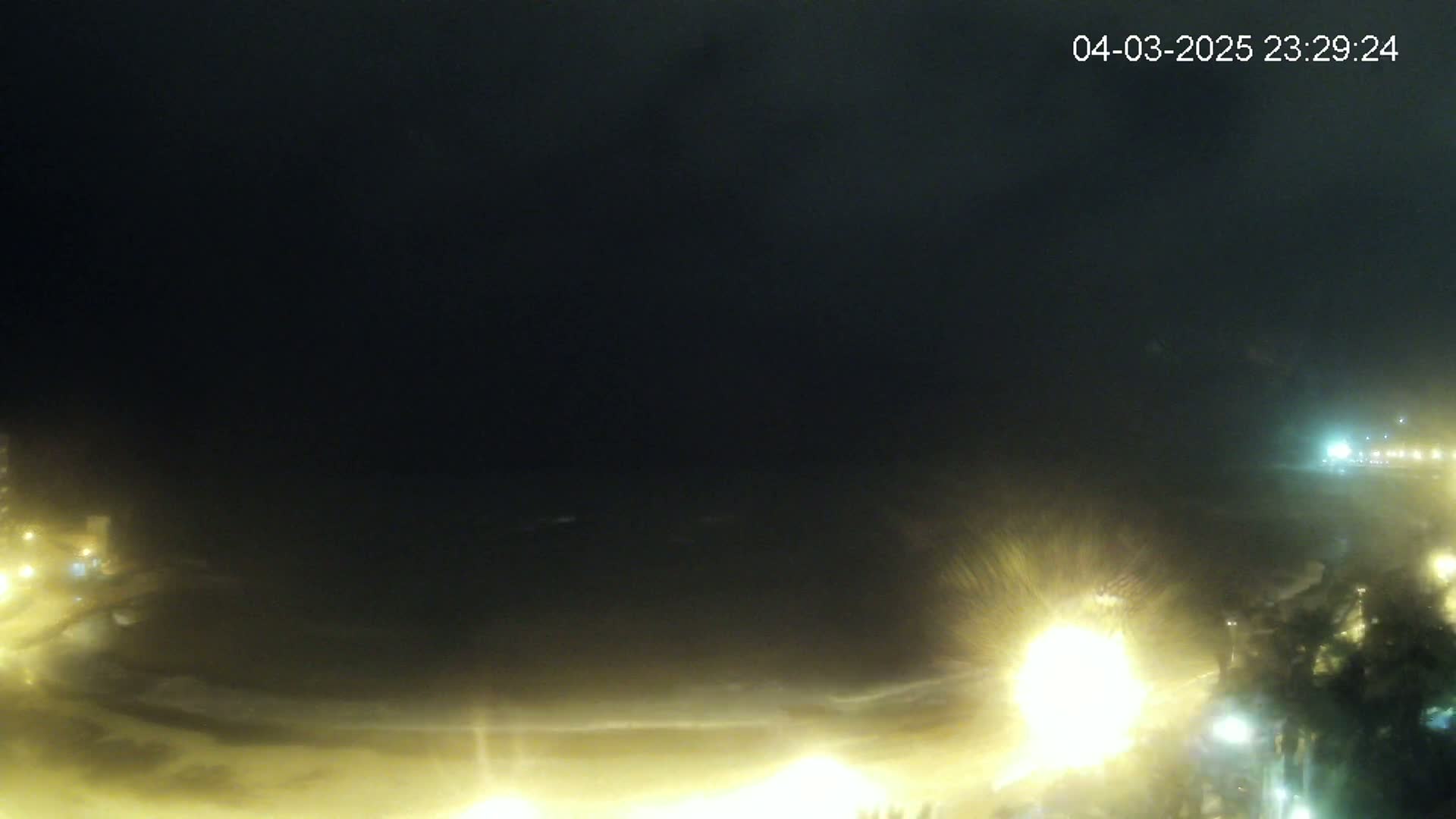 A nighttime view of a beach with dimly lit shoreline and dark, stormy ocean under a dark sky.