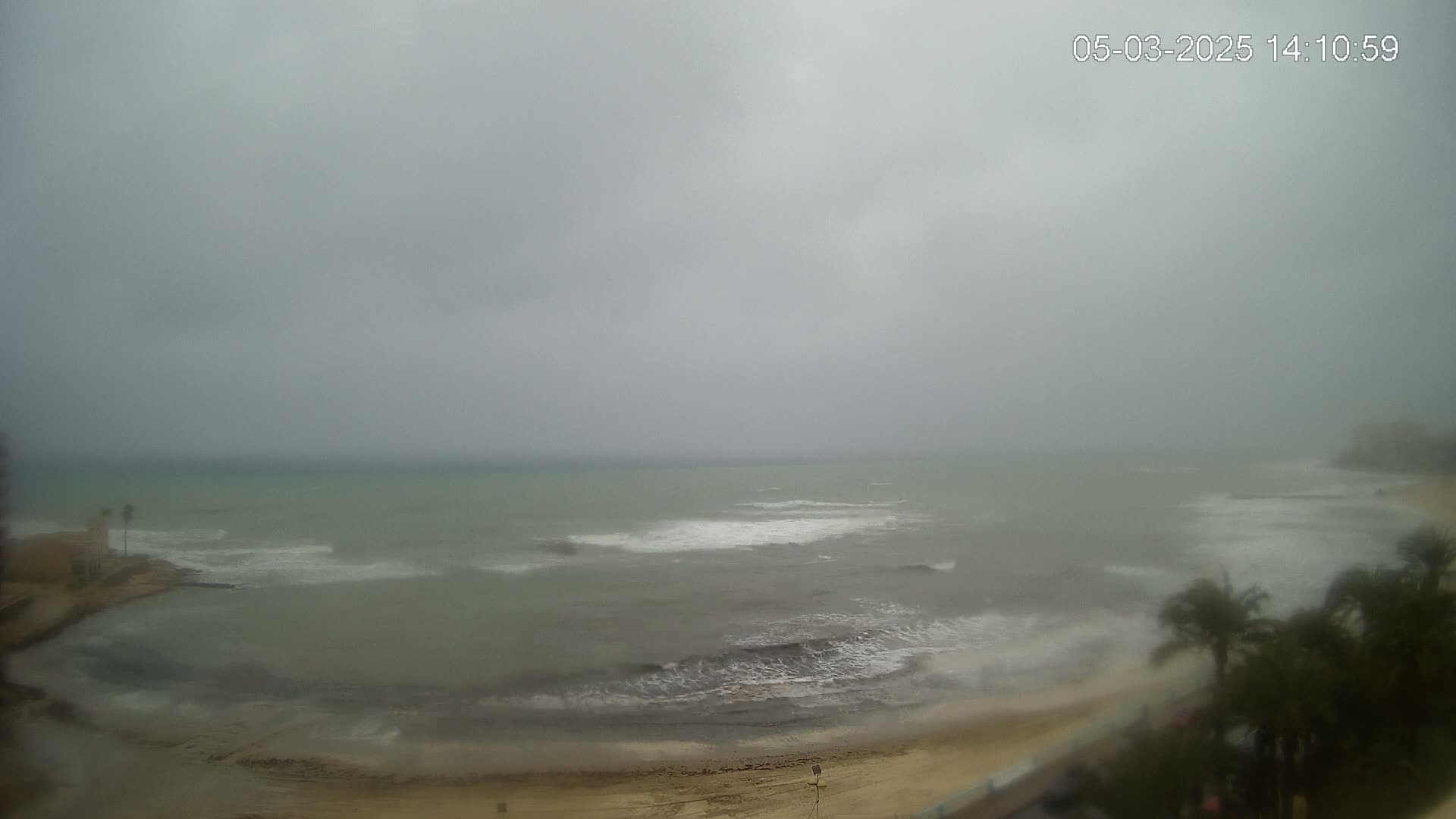 A hazy, overcast day shows a beach with moderate waves crashing onto the shore, with palm trees visible in the lower right corner.