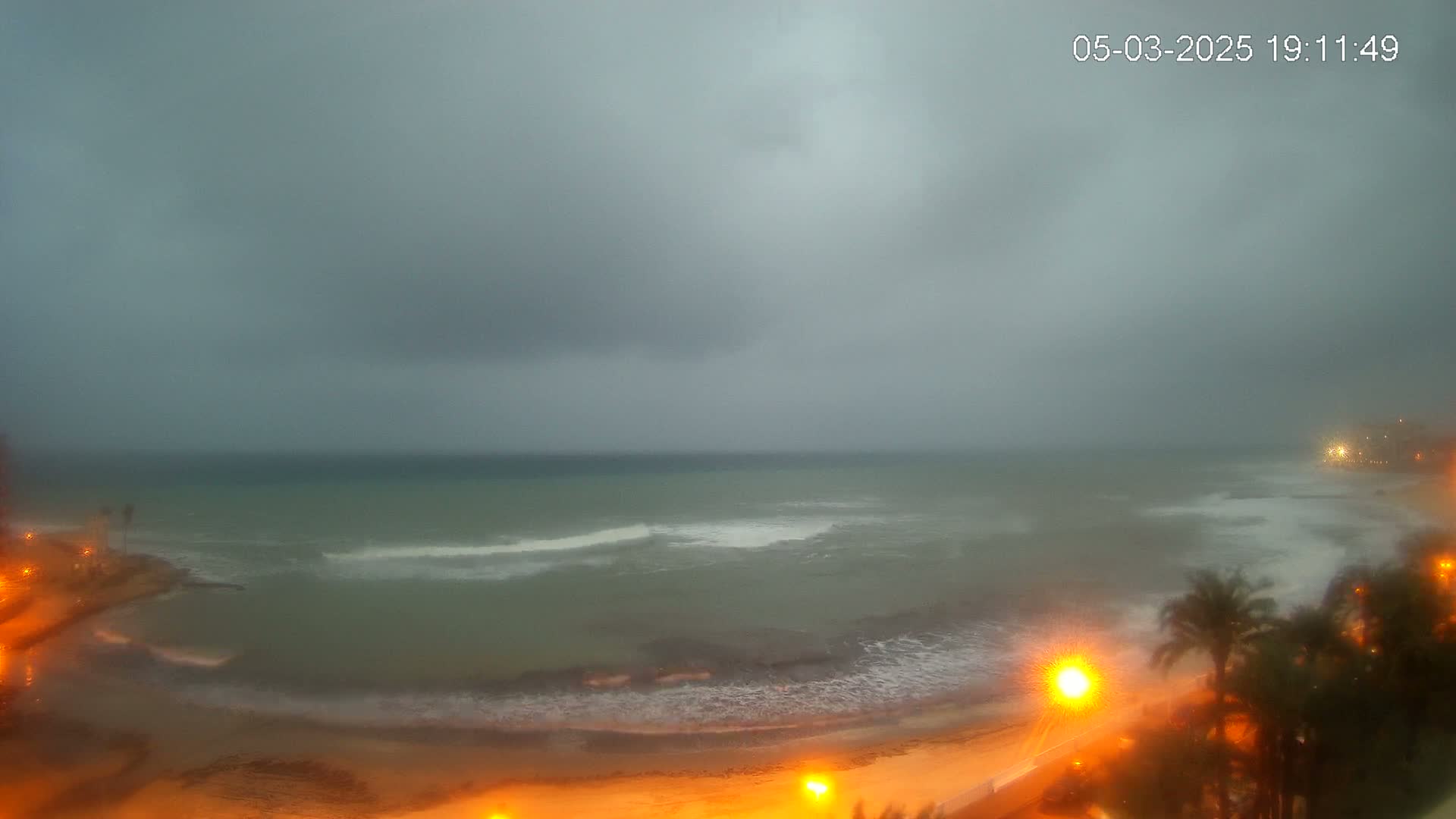 A beach at night under a cloudy, overcast sky shows moderate waves breaking on the shore, with palm trees and lights visible along the coast.
