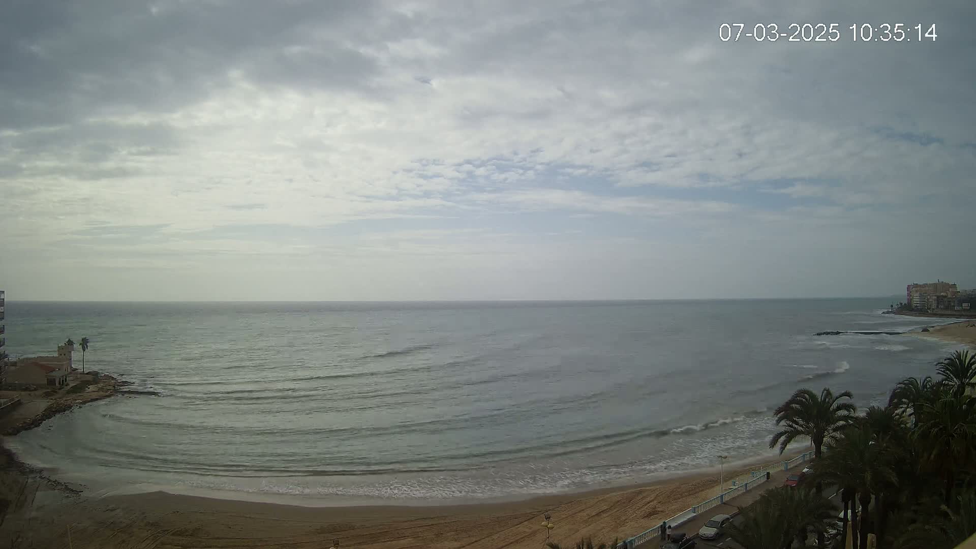 A mostly cloudy day shows a sandy beach with calm waves lapping the shore, palm trees, and buildings in the distance.