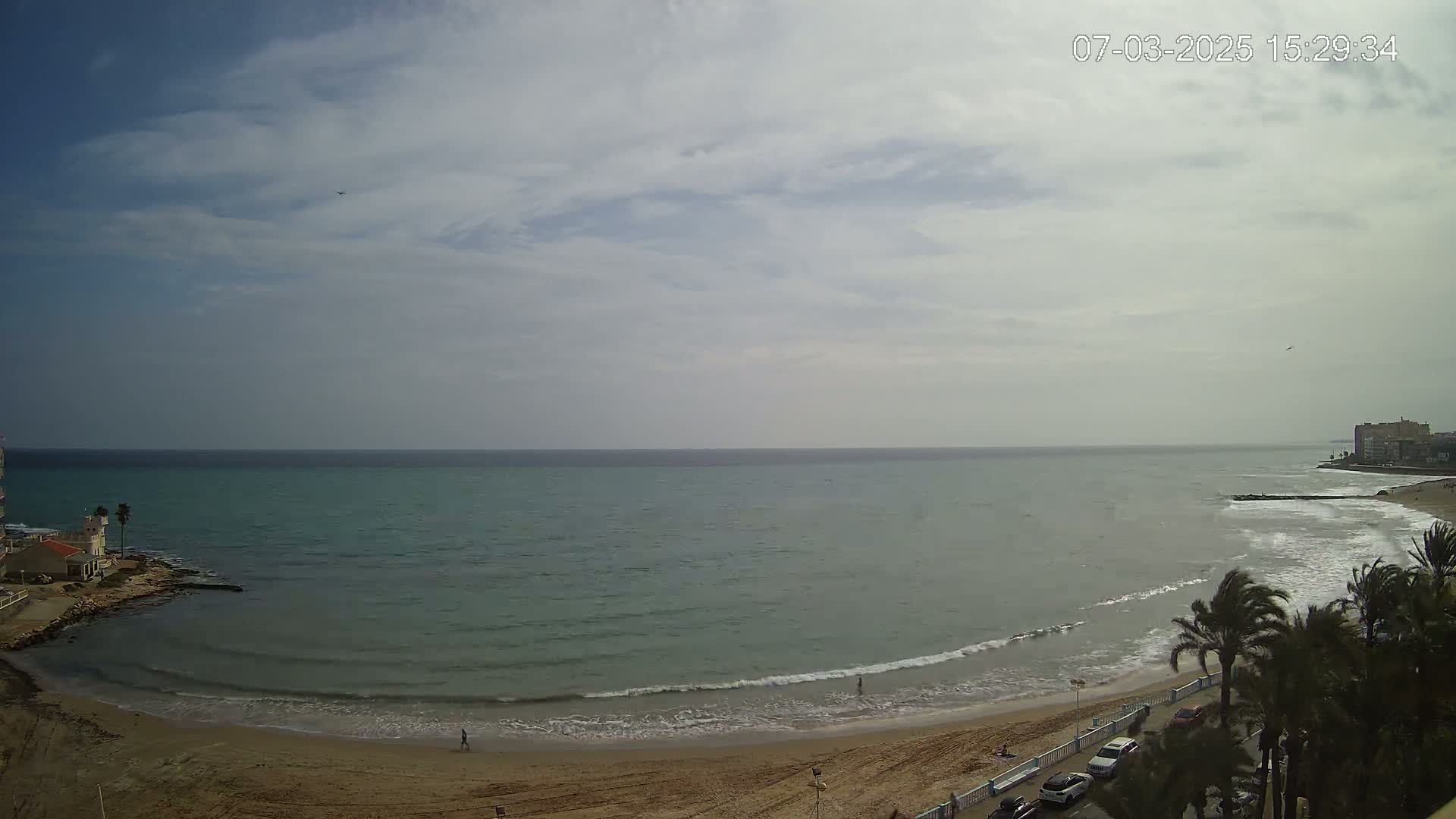 A sandy beach curves around a calm, teal sea under a mostly cloudy sky.