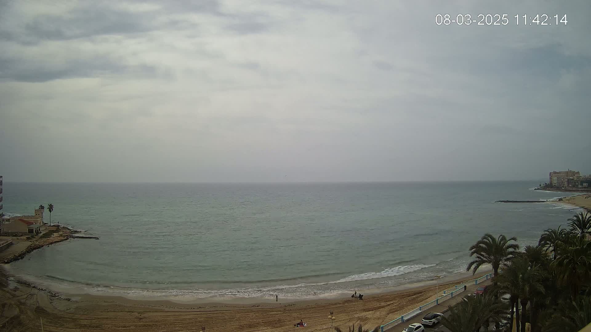 A mostly cloudy day shows a sandy beach curving into a calm sea with small waves, bordered by palm trees and buildings.