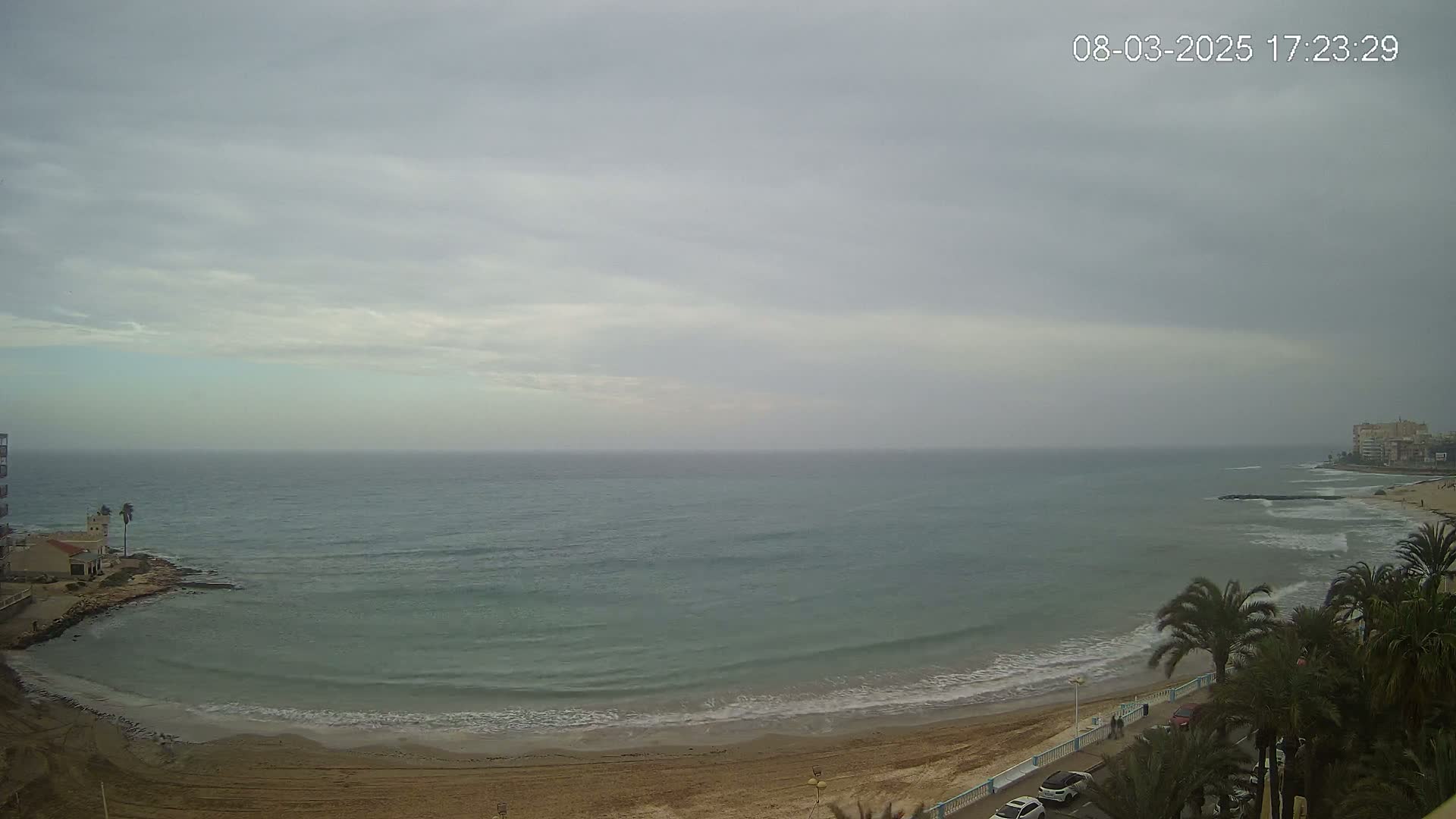 A sandy beach with calm waves under an overcast sky meets a calm sea next to buildings and palm trees.