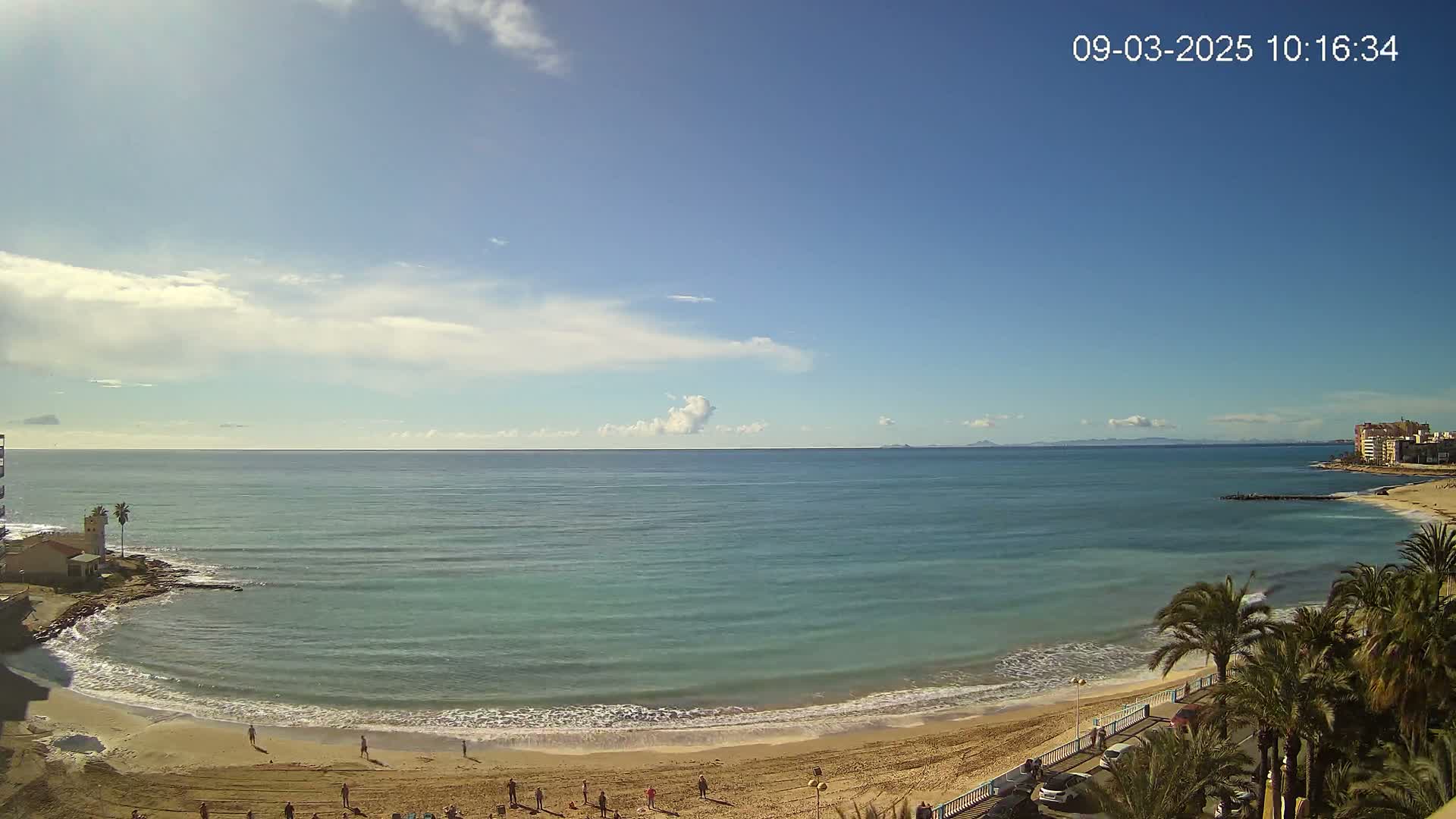 A sandy beach curves along a calm, teal ocean under a mostly clear, bright blue sky with a few scattered clouds.
