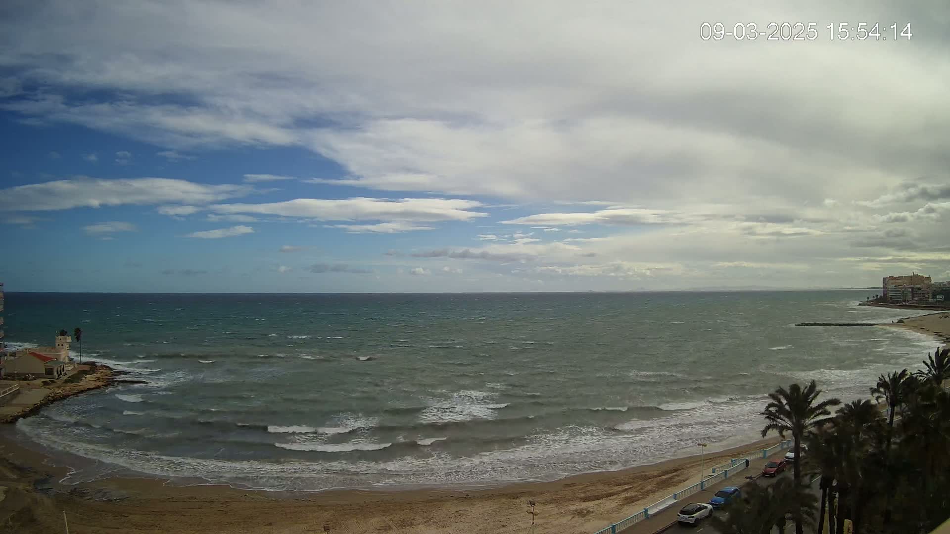 A windy day with partly cloudy skies shows a wide expanse of choppy ocean waves meeting a sandy beach lined with palm trees and a few cars.