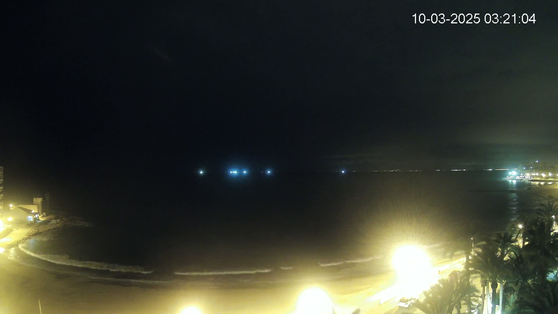 A nighttime view of a beach with gently lapping waves, illuminated by streetlights, under a dark, clear sky.