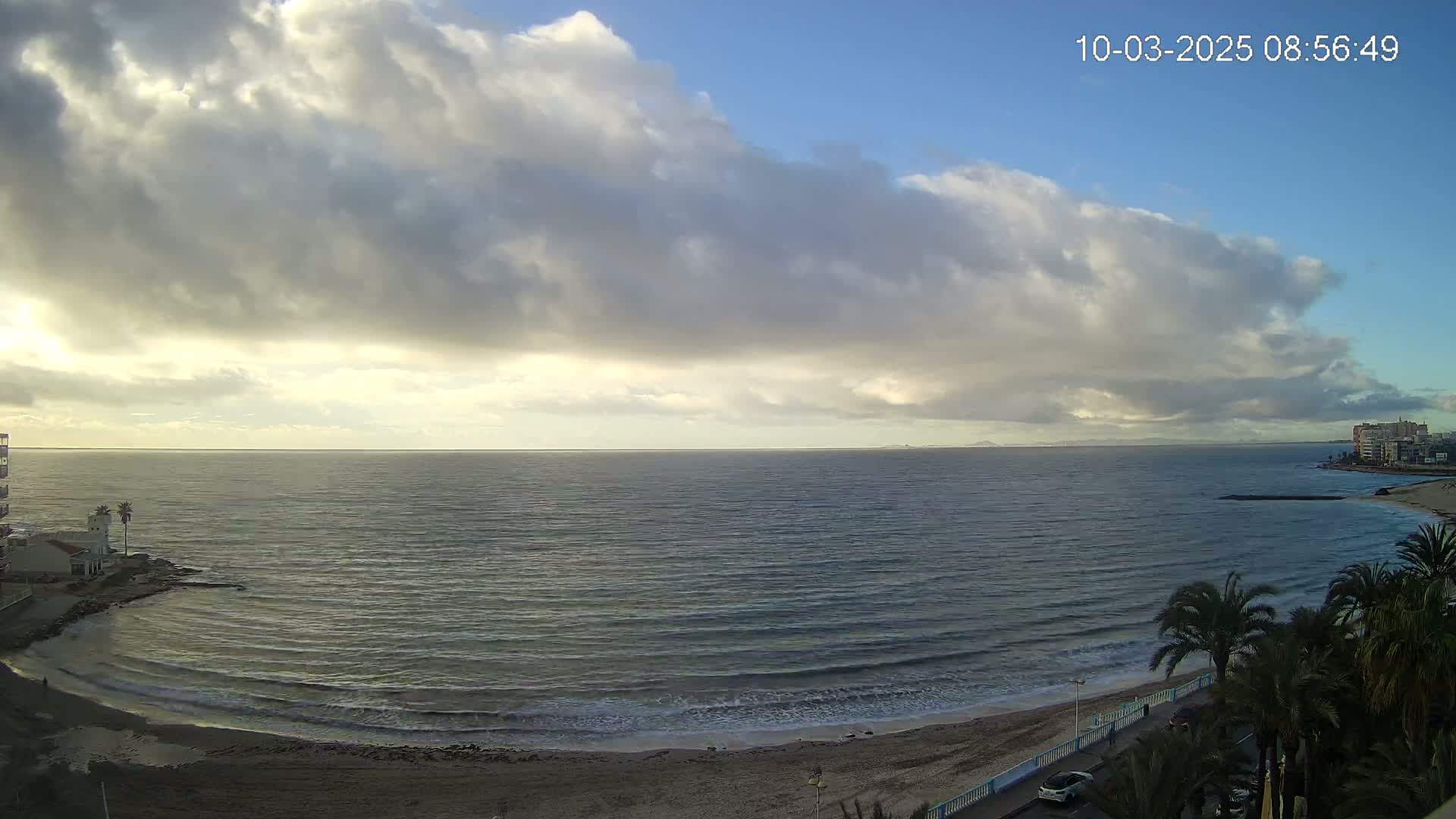 A mostly cloudy day reveals a calm sea with gentle waves lapping a sandy beach lined with palm trees and buildings.