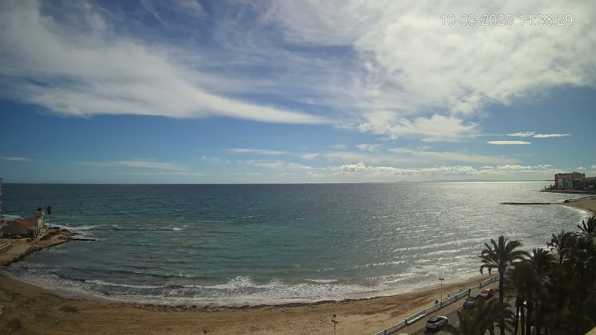 A partly cloudy day shows a sandy beach with palm trees, calm waves lapping the shore, and a calm sea extending to a distant coastline under a mostly blue sky.