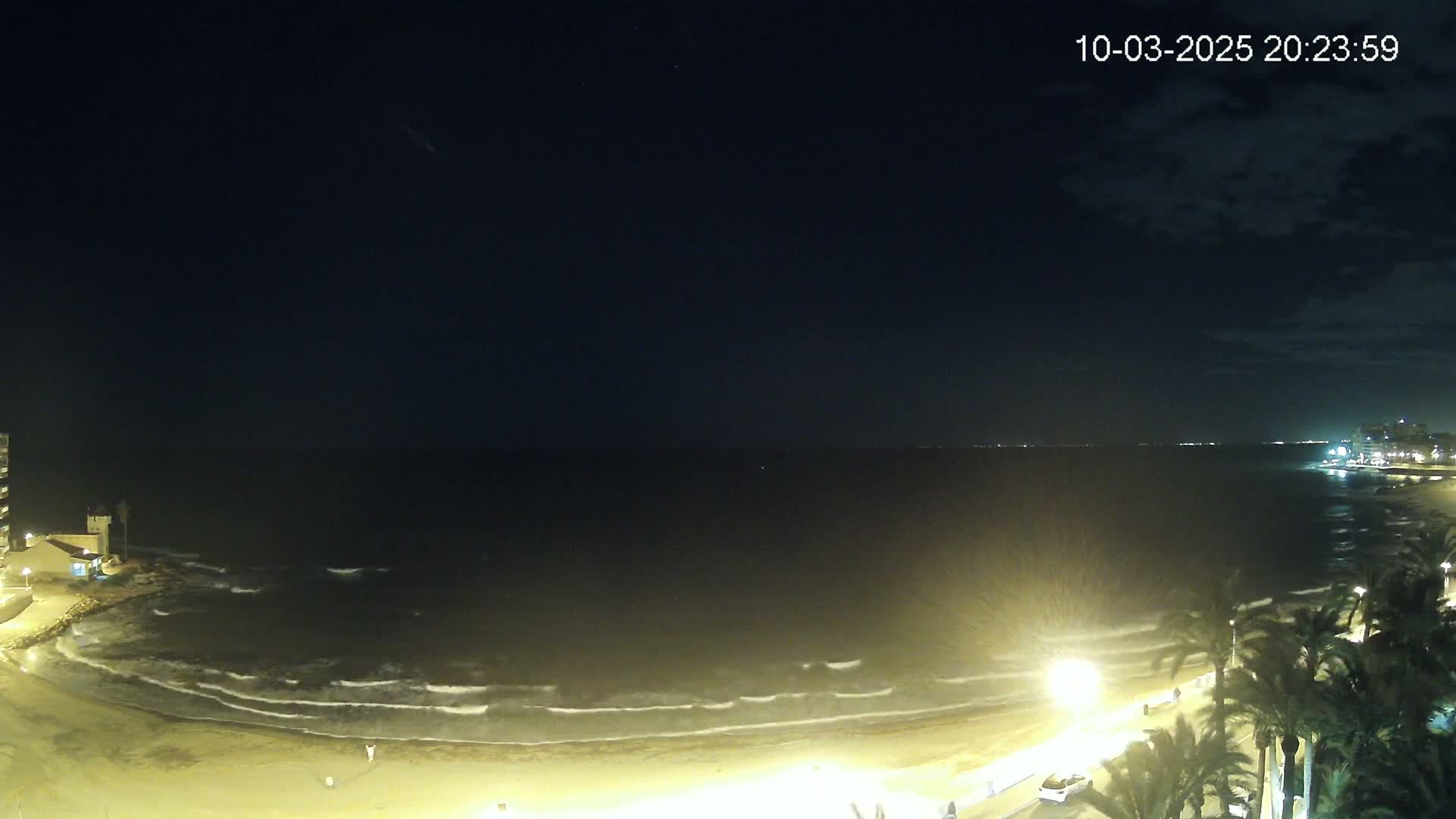 A nighttime view of a sandy beach with gentle waves lapping the shore, under a mostly clear sky with a few faint clouds.