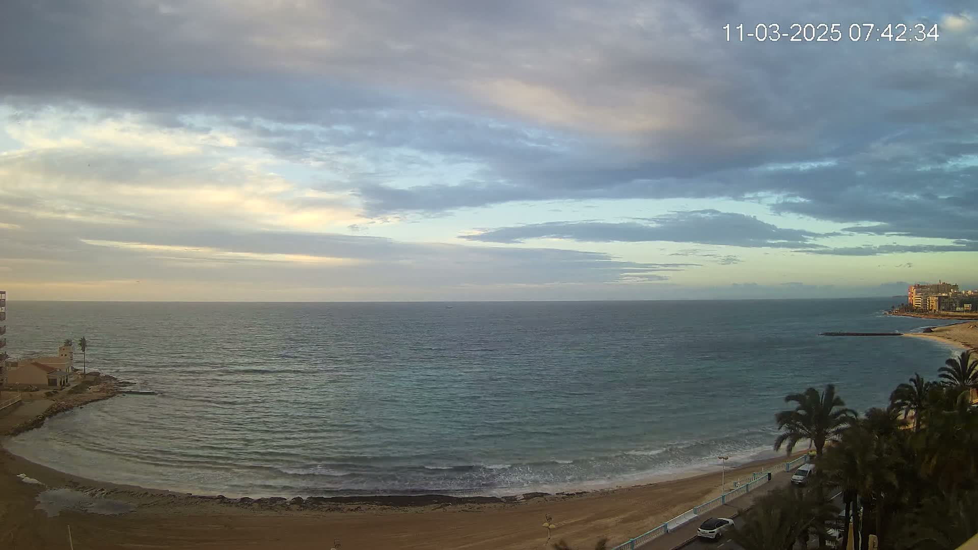 A mostly cloudy sunrise over a calm sea, with a sandy beach and palm trees visible in the foreground.
