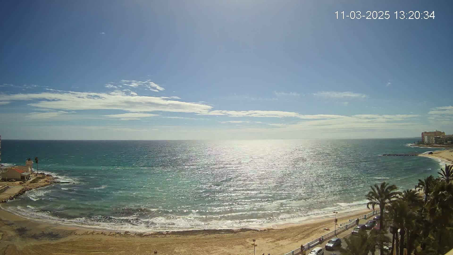 A sunny day with a partly cloudy sky shows a view of a sandy beach, ocean waves, and palm trees lining a road next to the beach.