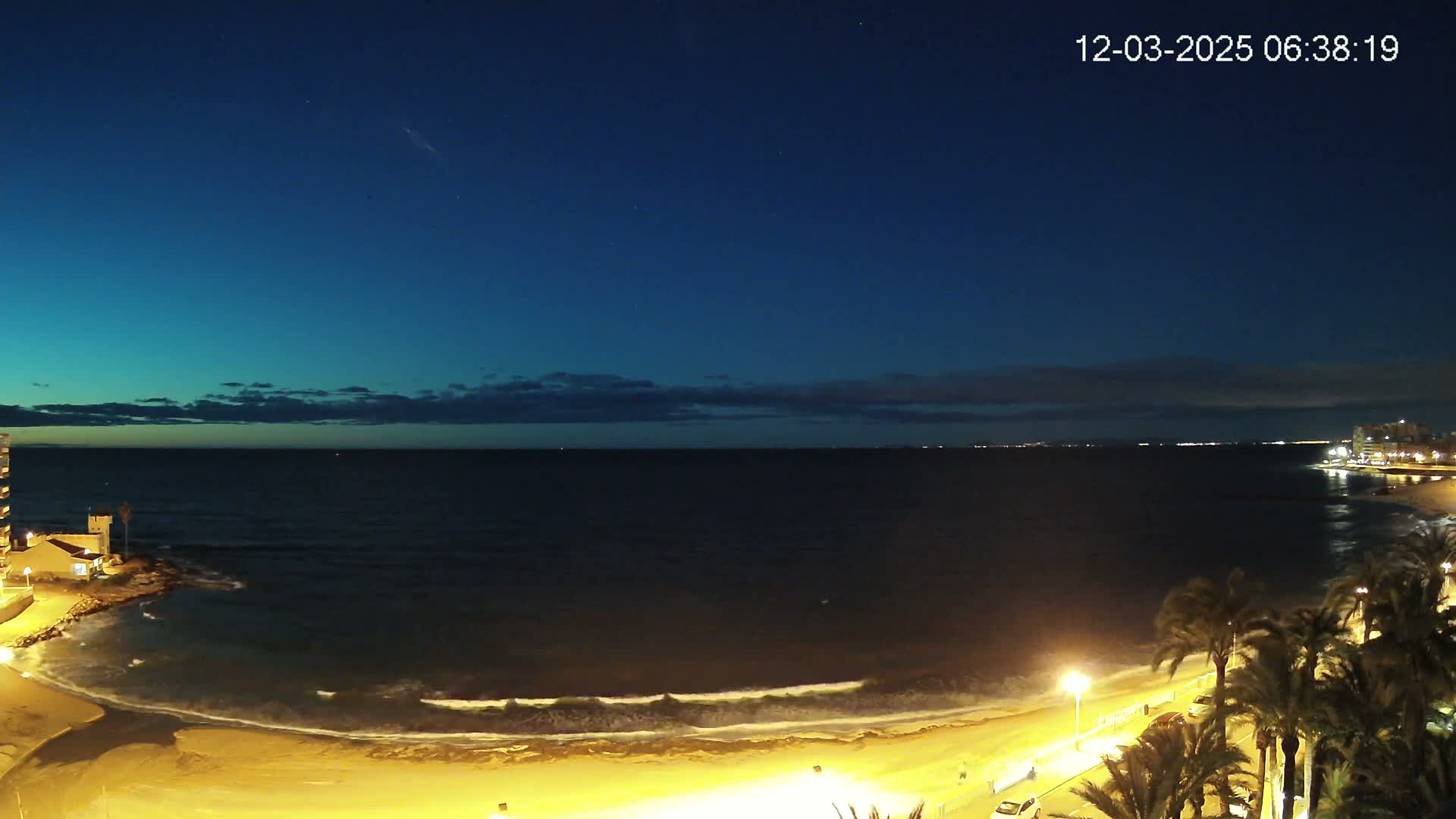 A dark blue night sky over a gently lapping, lit shoreline with palm trees and buildings along the coast.