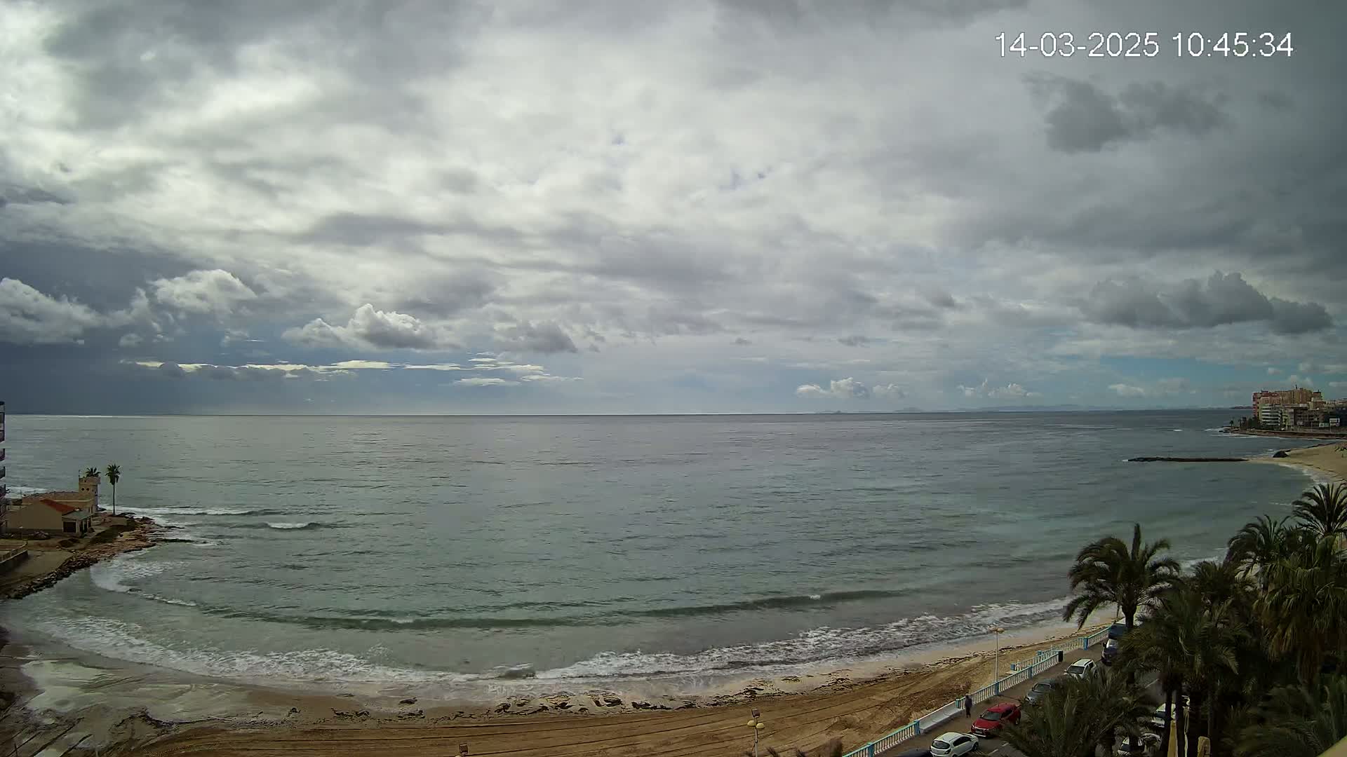 A mostly cloudy day overlooks a calm sea with gentle waves lapping a sandy beach lined with palm trees and a roadway.
