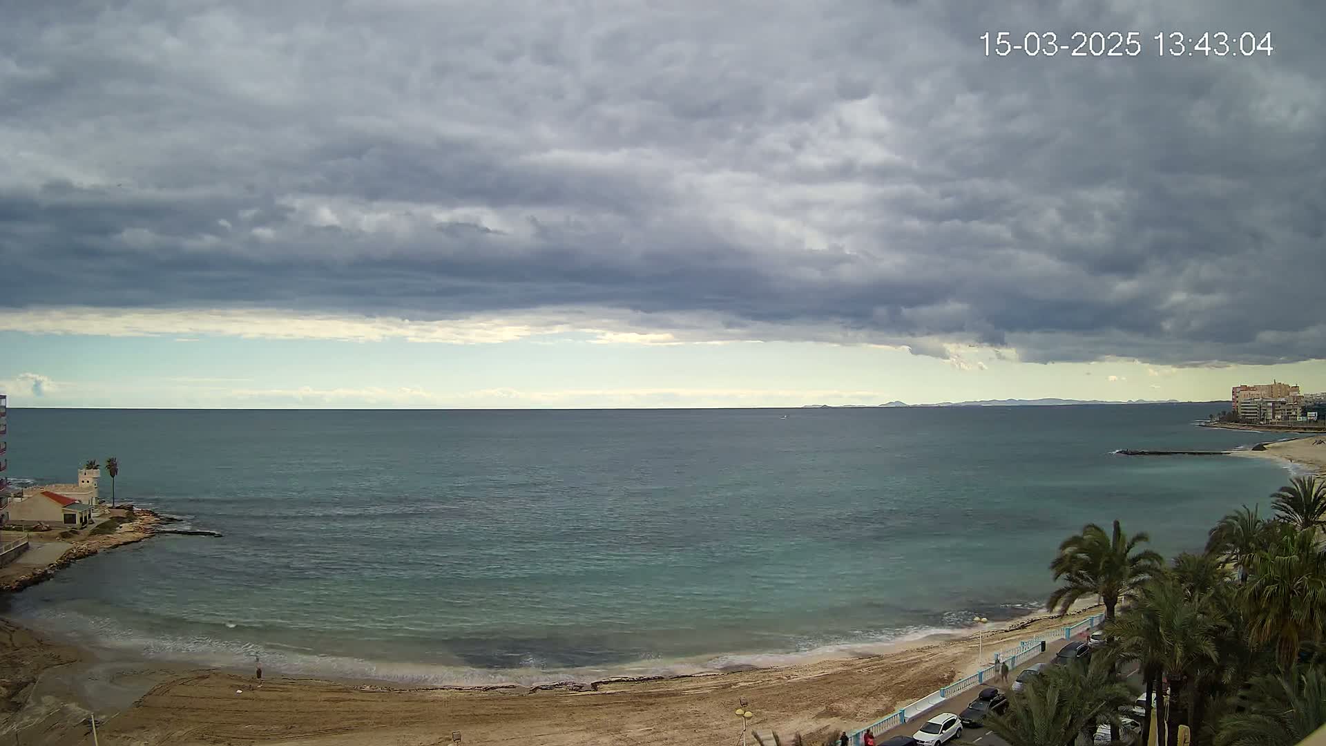 A sandy beach with palm trees and buildings borders a calm, teal sea under a mostly cloudy, gray sky.