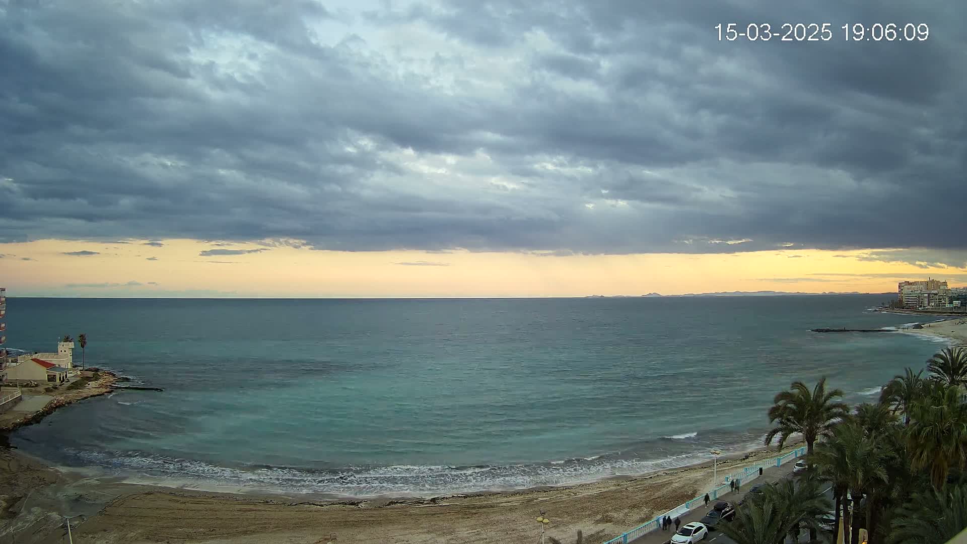 A sandy beach with palm trees lines a calm, teal ocean under a mostly cloudy, dusky sky.