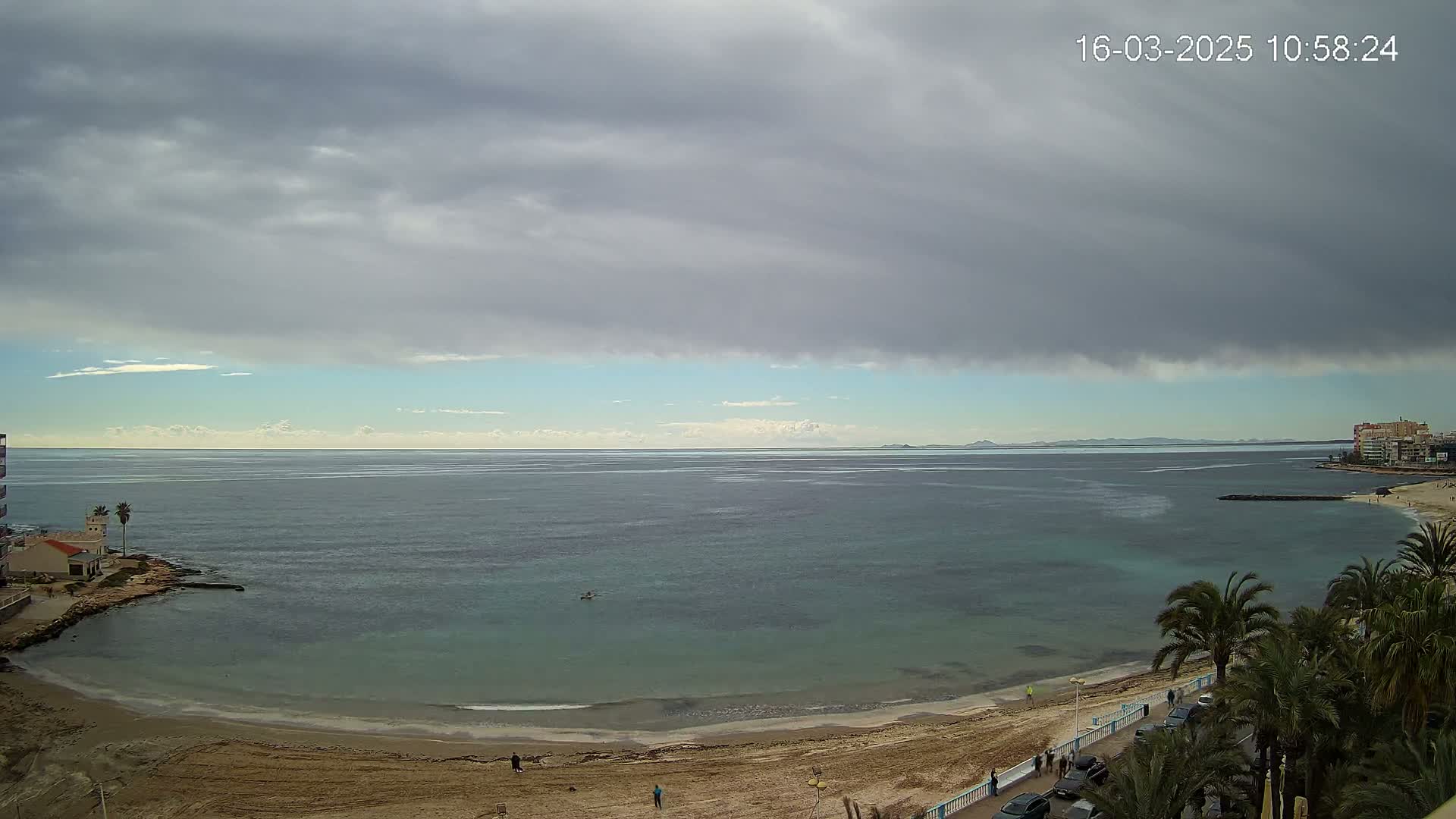 A mostly cloudy day overlooks a calm, teal sea lapping a sandy beach lined with palm trees and buildings.