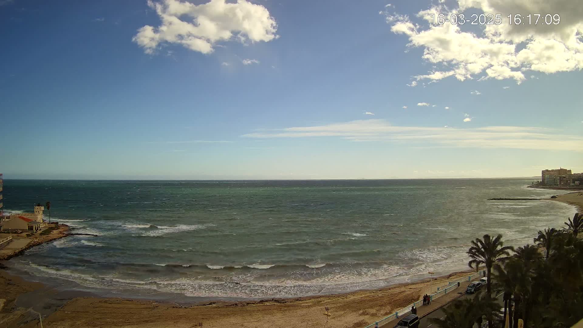 A windy, partly cloudy day at a beach with a sandy shore, waves crashing, and palm trees lining a coastal road.